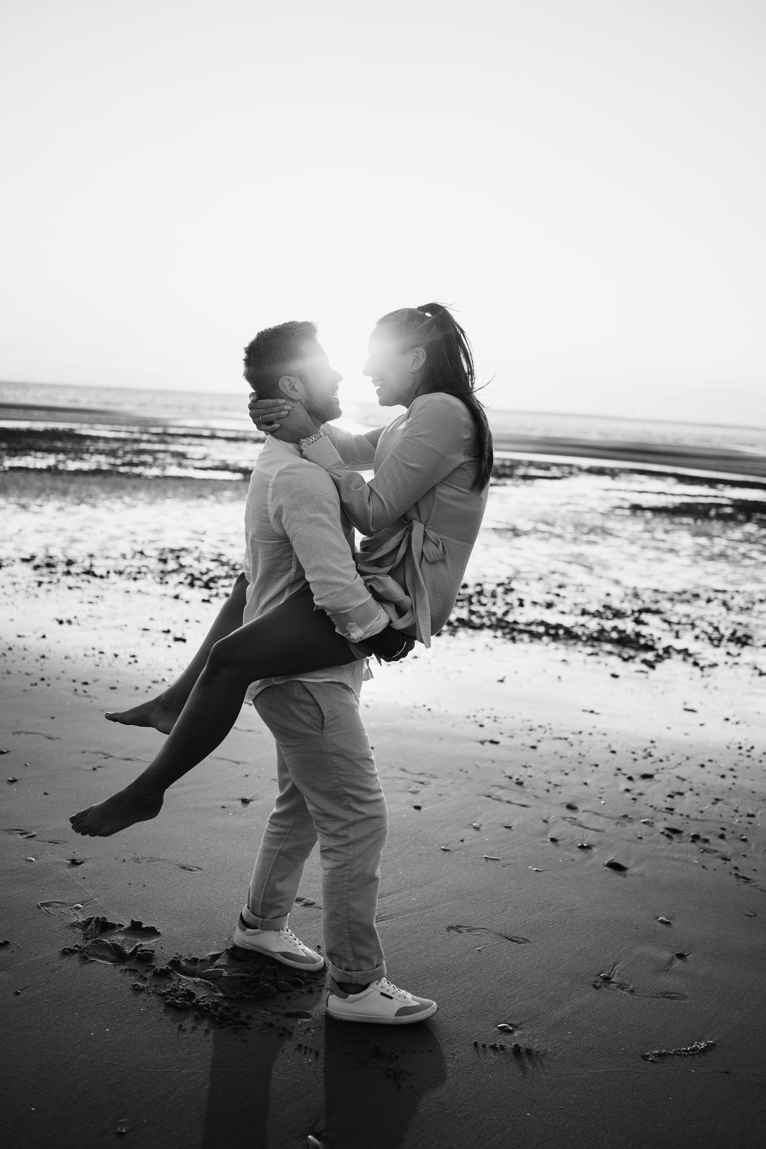 Black and white photo of a couple embracing on a beach at sunset, man lifting woman as they lean in for a kiss.