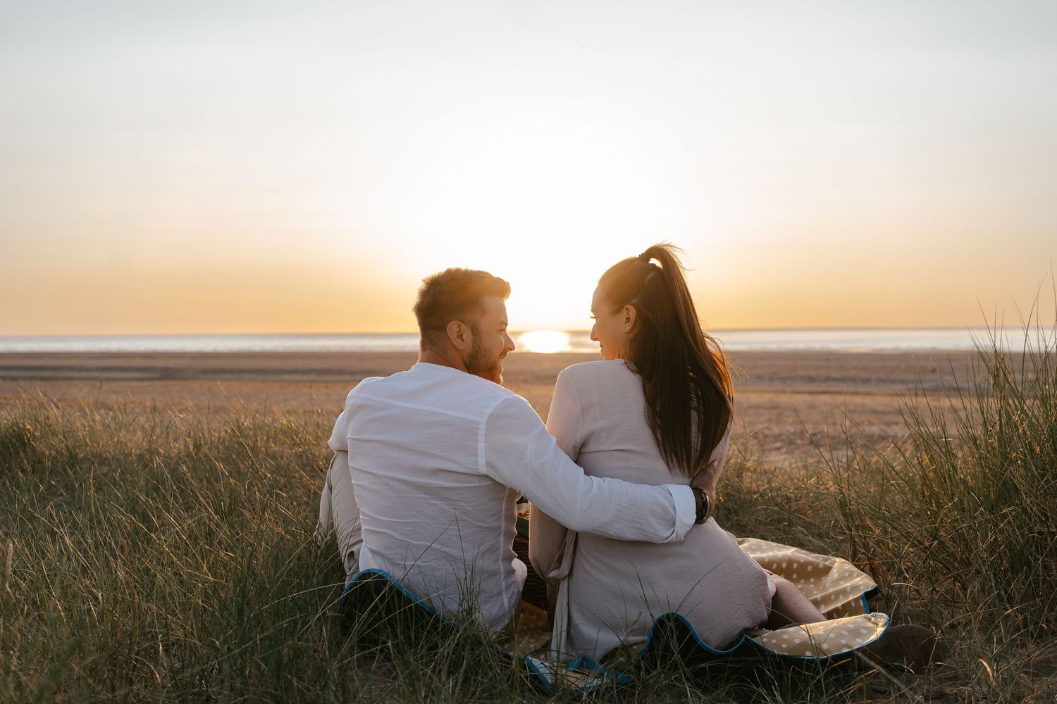 Couple sitting together in beach grass watching a stunning golden sunset over the shore.