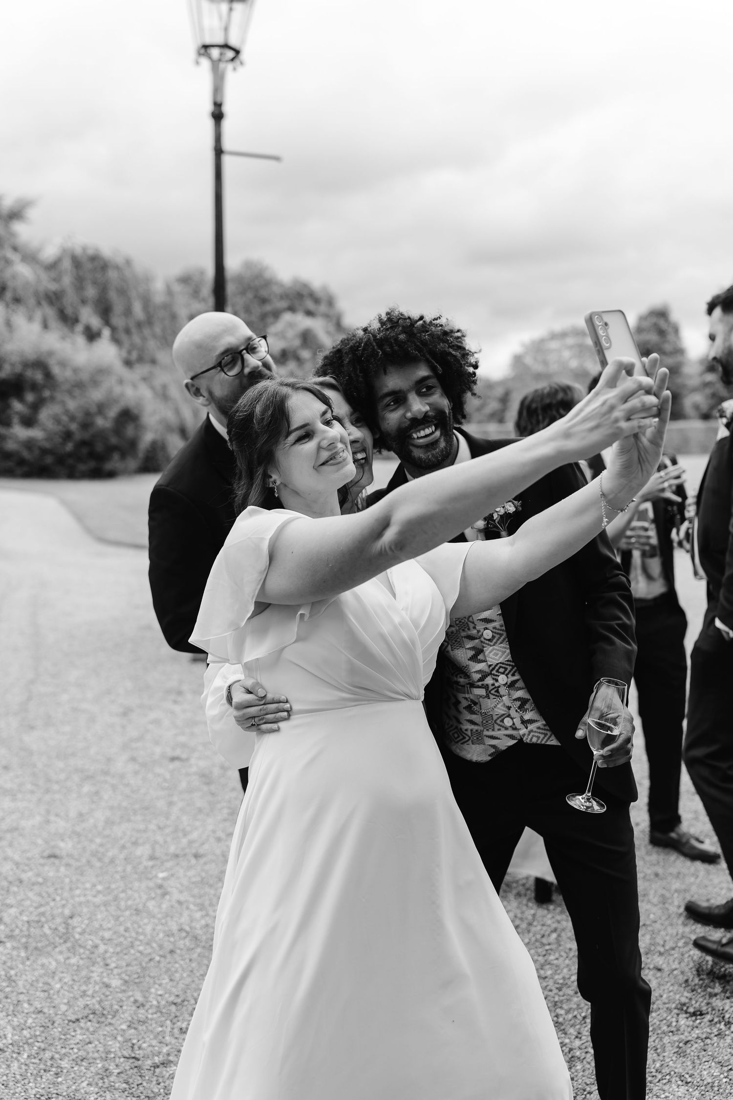 Bride takes a selfie with smiling wedding guests outdoors in black and white photo.