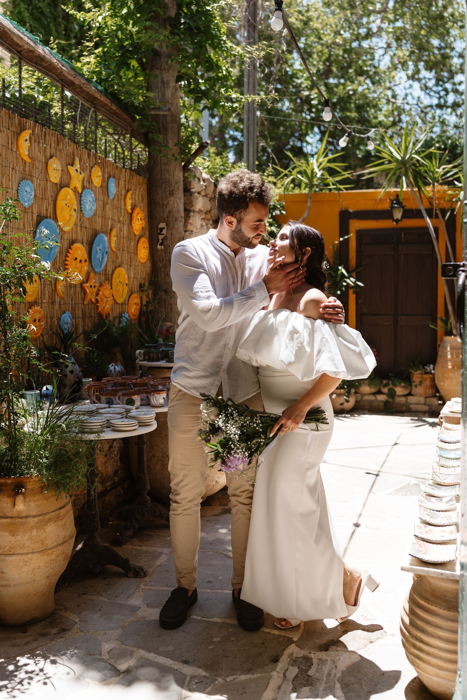 Groom kissing bride in sunny garden courtyard surrounded by colorful pottery and lush green plants.