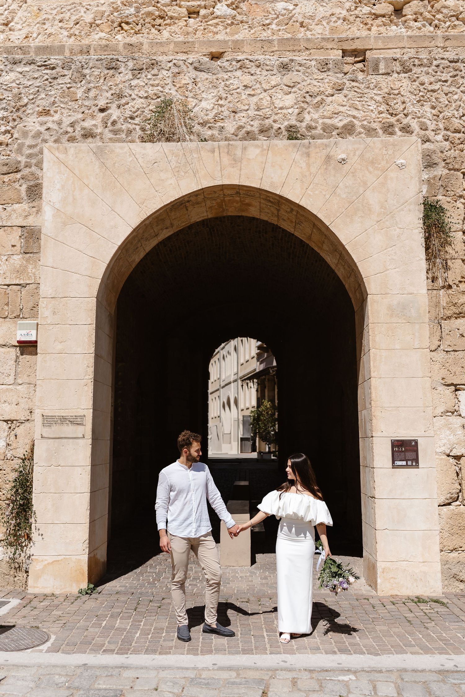 Couple holding hands under a stone archway in an ancient Mediterranean old town on a sunny day.