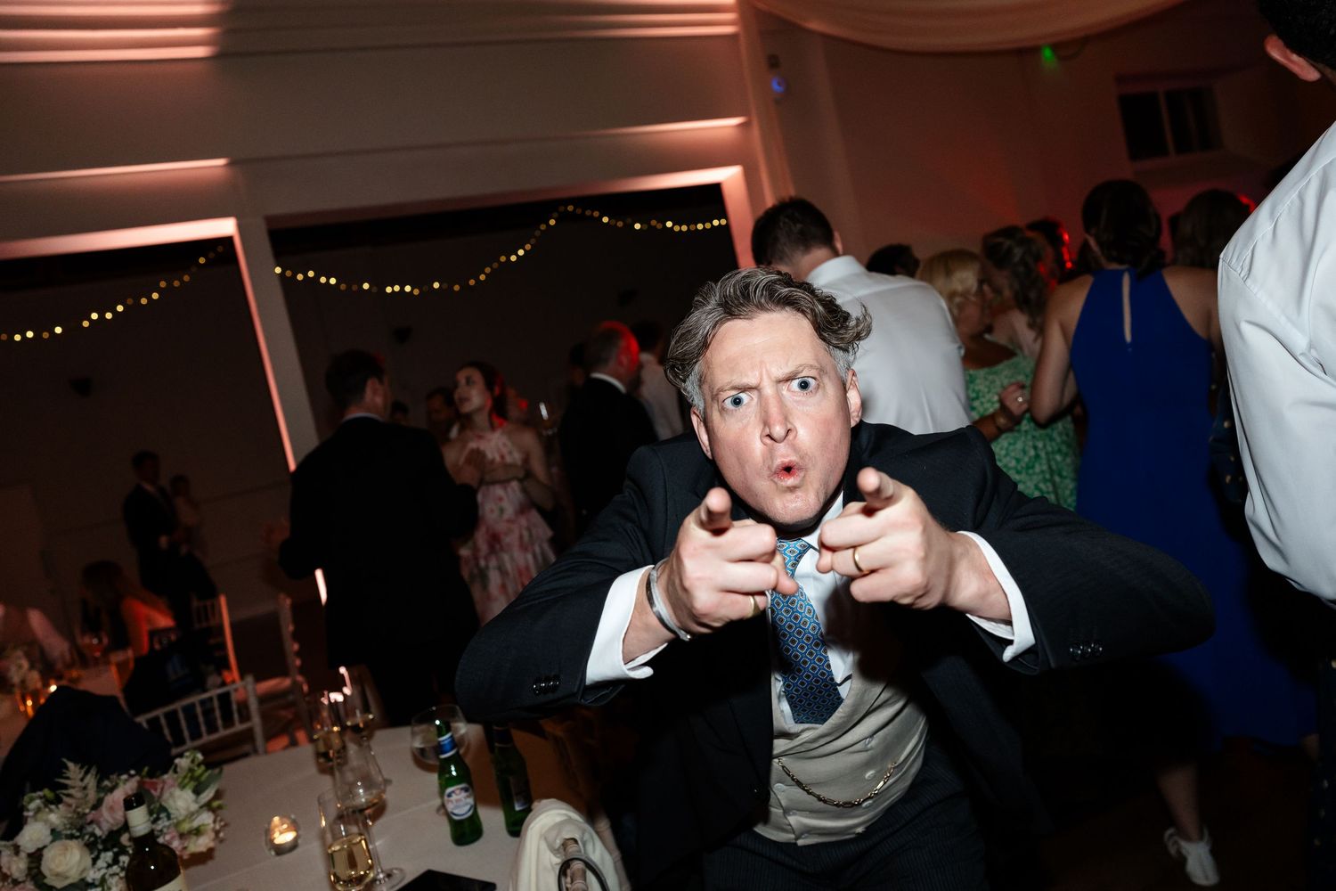 Man in black tuxedo pointing at camera with surprised expression at elegant wedding reception venue.