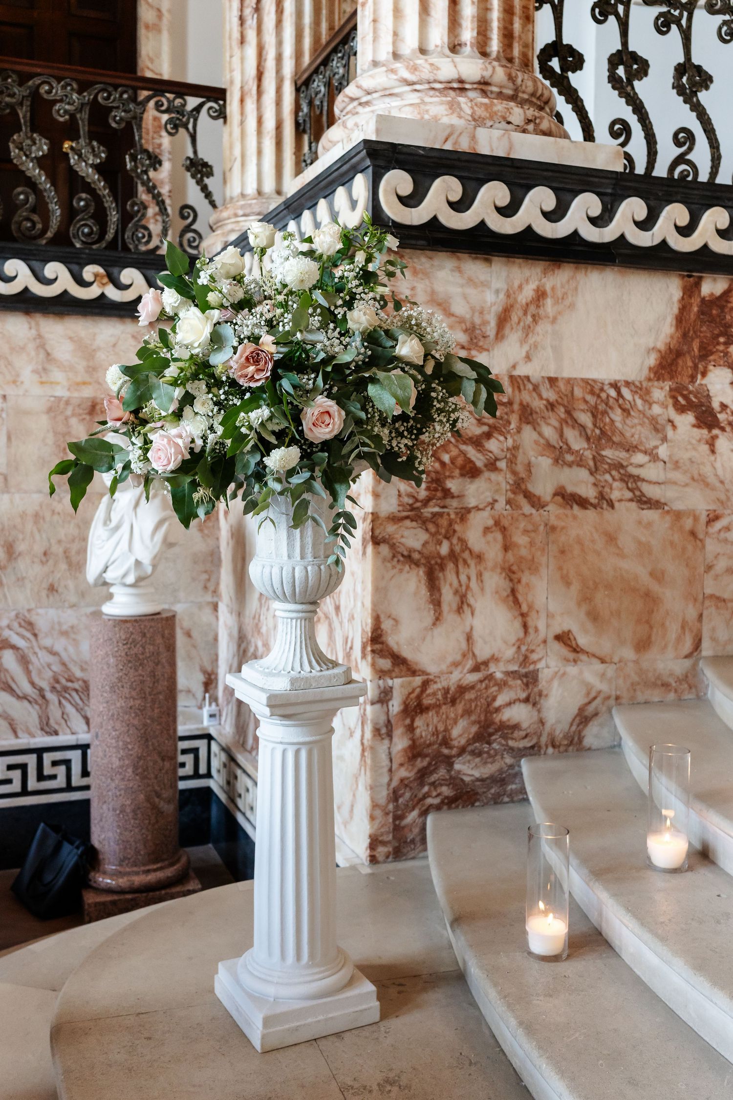 Elegant white urn floral arrangement with roses and greenery on a pedestal beside marble stairs with candles.