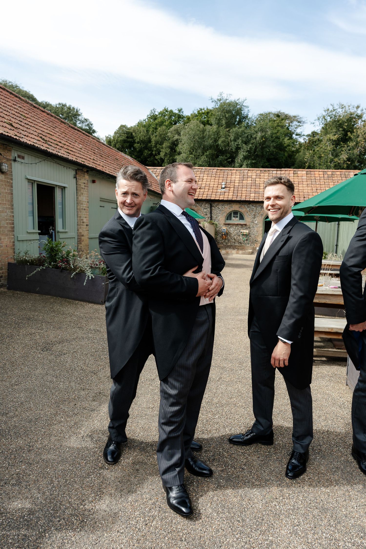 Three groomsmen in black suits laughing and joking together outside a rustic wedding venue on a sunny day.
