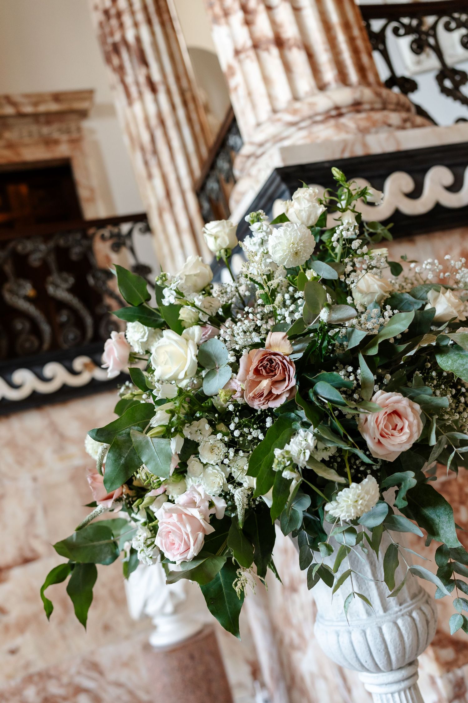 Elegant wedding floral arrangement with pink and white roses in a white urn vase, set in a marble interior.