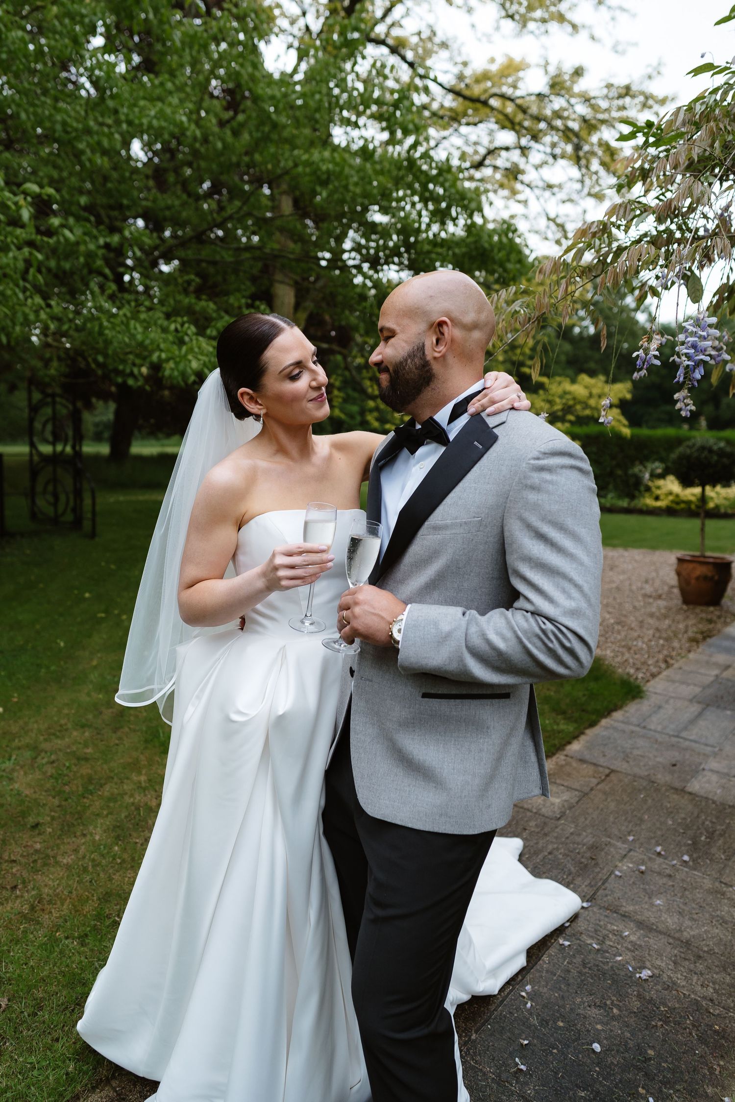 Elegant bride and bald groom share a romantic moment in a lush garden holding champagne glasses.