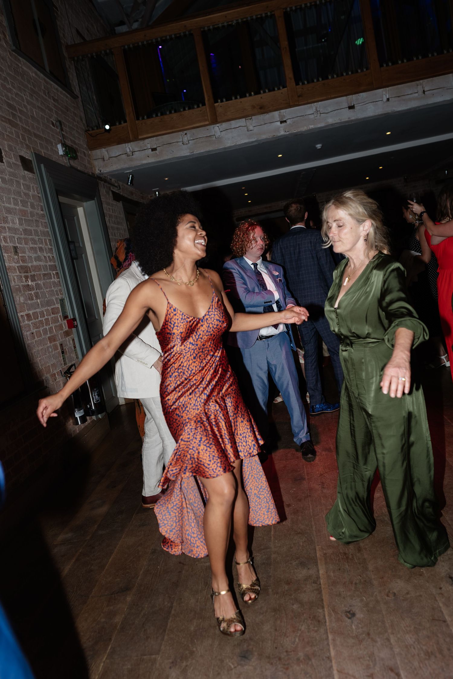 Two women in elegant dresses dance together joyfully at a wedding reception.