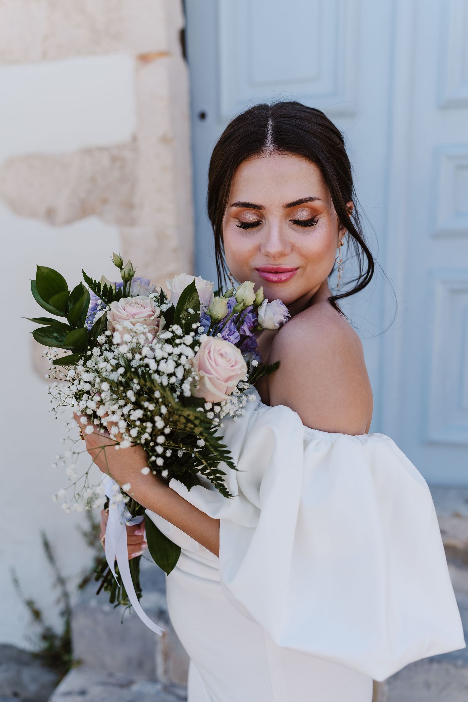 Beautiful dark-haired bride in off-shoulder white gown smells her pastel rose and wildflower bouquet.