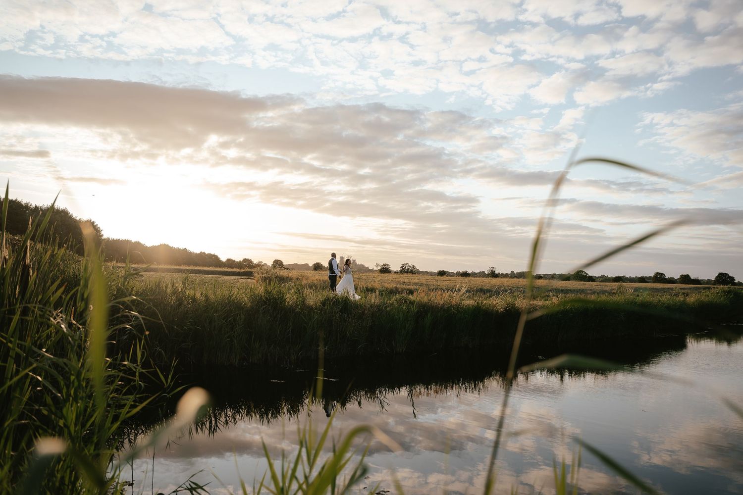 Couple standing by a reflective pond in a lush green field at golden hour sunset.
