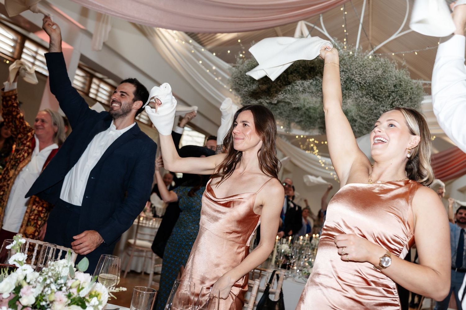 Joyful wedding guests dancing and waving napkins in elegant attire at a decorated reception venue.