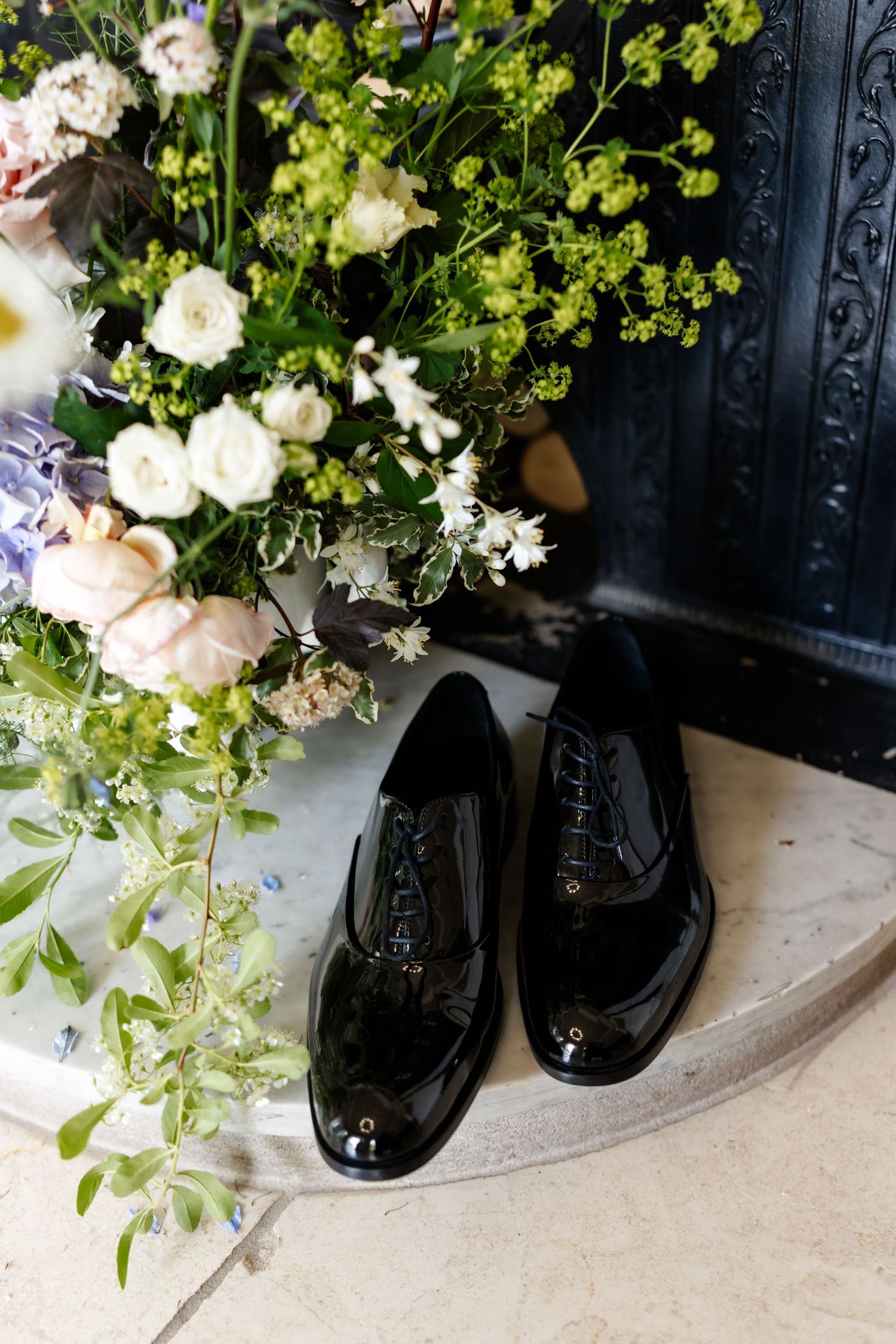 Shiny black patent leather Oxford shoes beside white and green floral arrangement on marble surface.