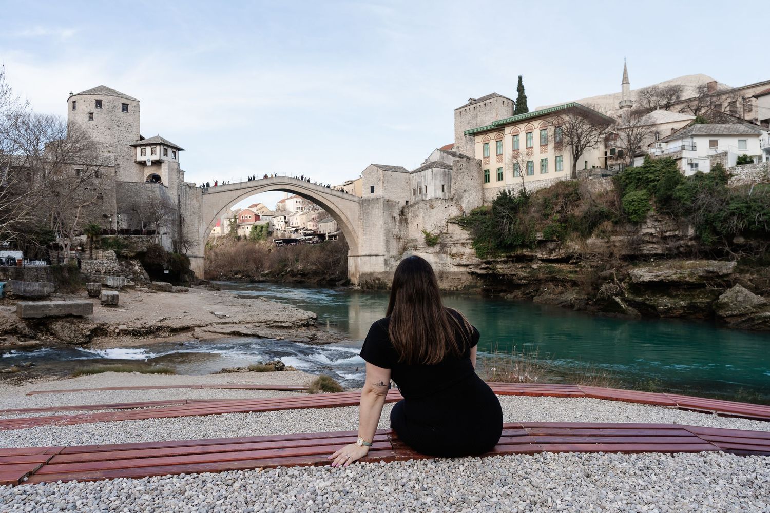 Woman sitting on a blanket admiring the historic Stari Most bridge over the turquoise Neretva River in Mostar.