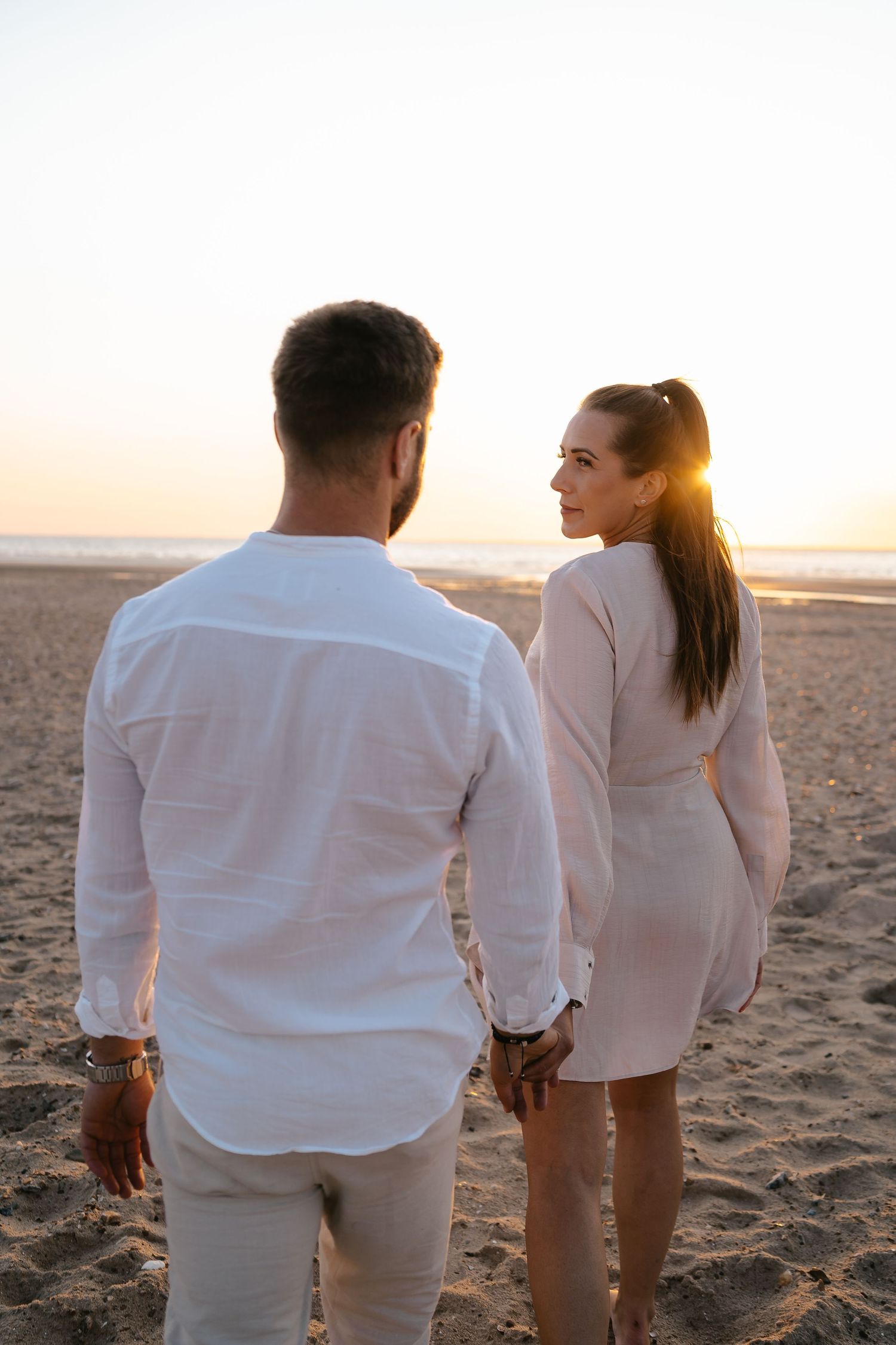 Couple holding hands on beach at golden sunset, woman looking back over shoulder.
