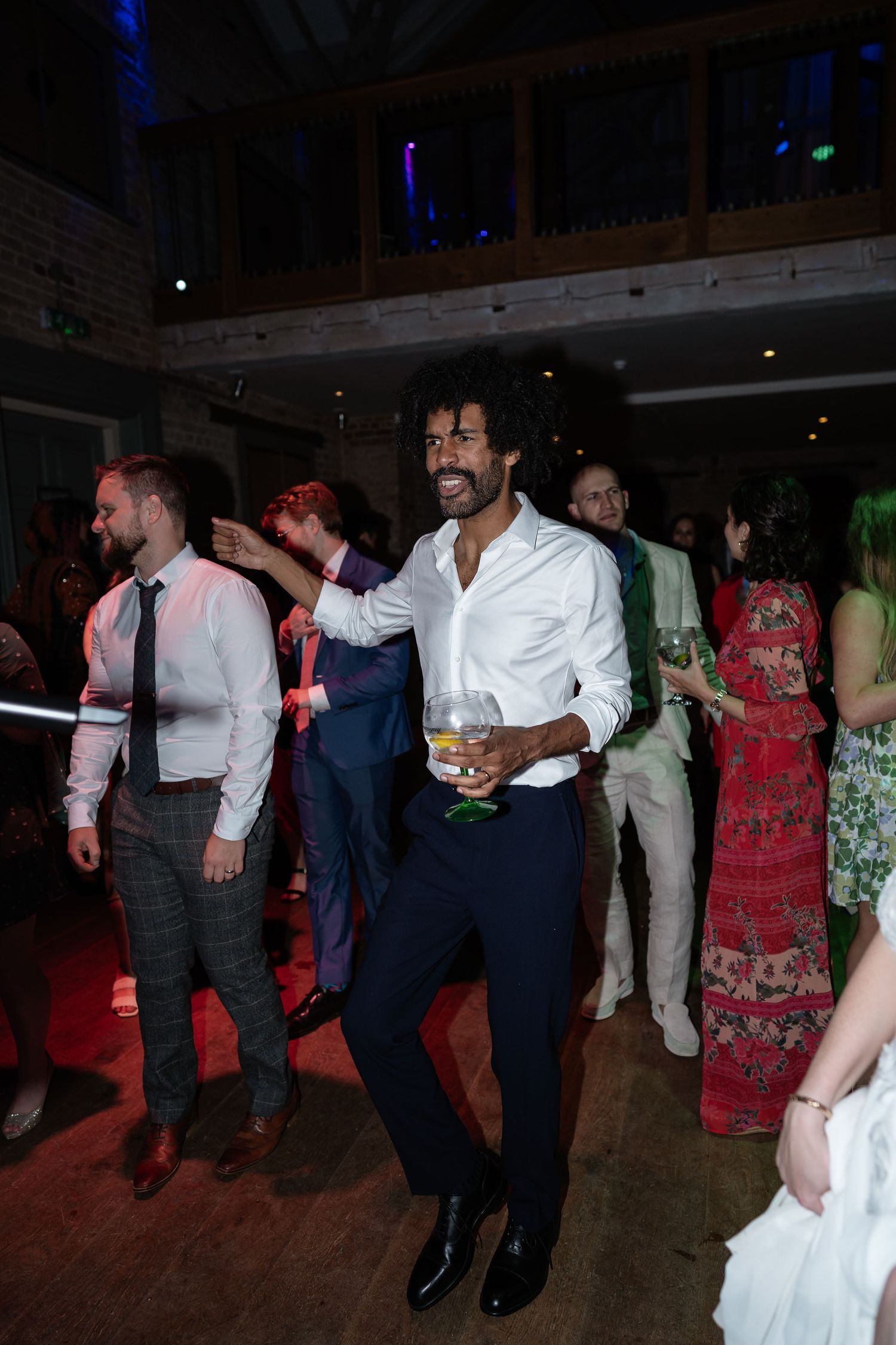 A stylish man in a white shirt dances with a drink at a lively wedding reception.