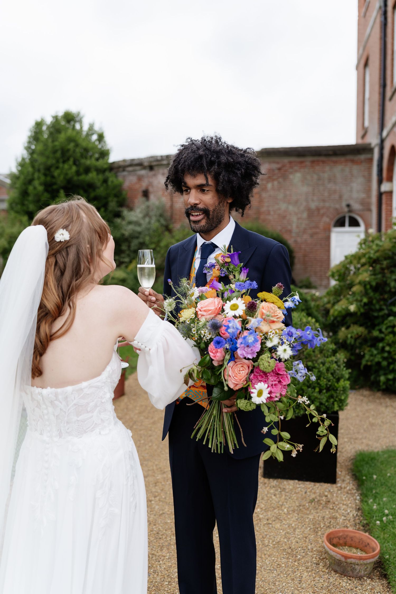 Smiling groom holds vibrant mixed flower bouquet as bride toasts with champagne outside brick venue.