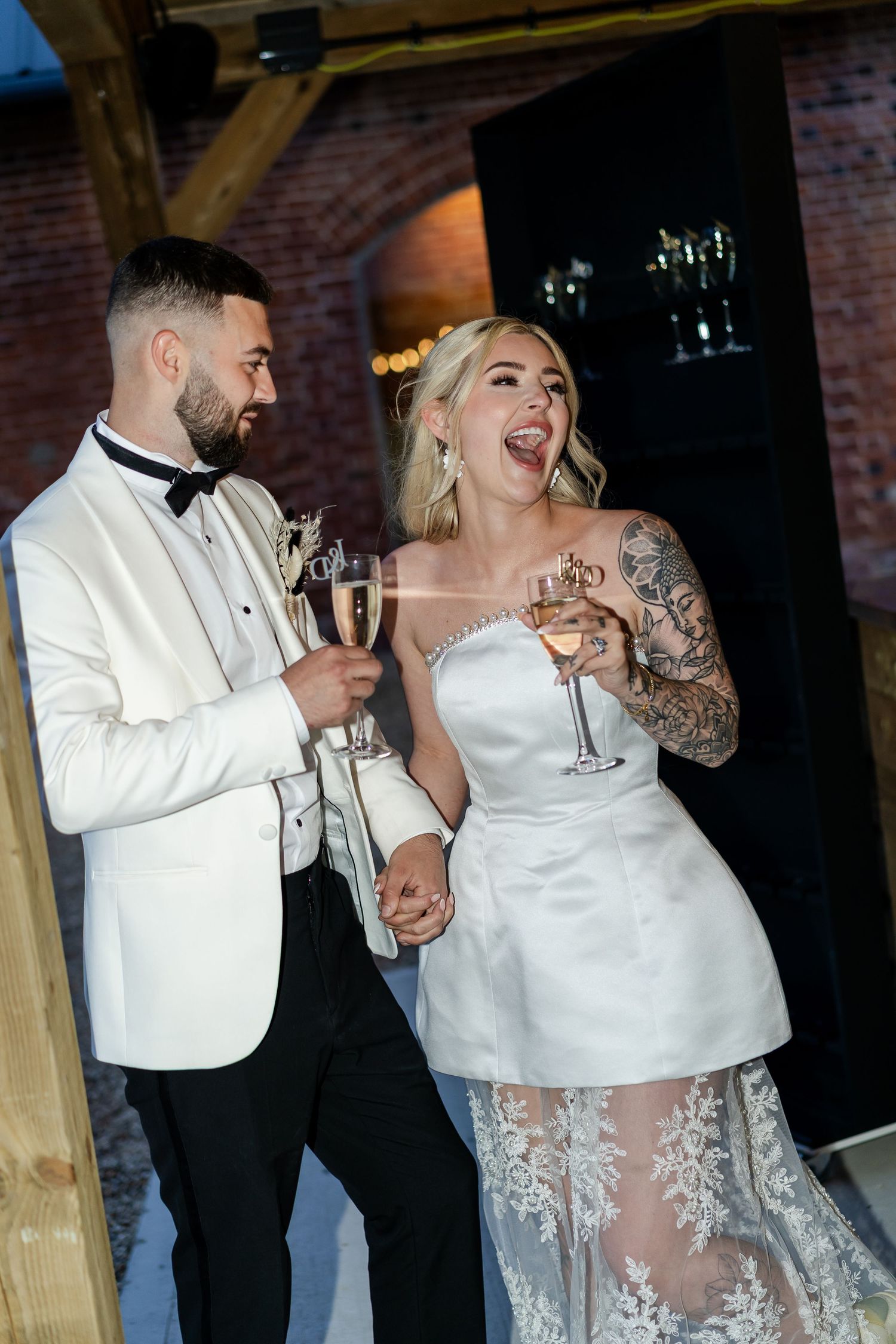 Bride with tattoos and groom in white tuxedo toast champagne glasses at wedding reception.
