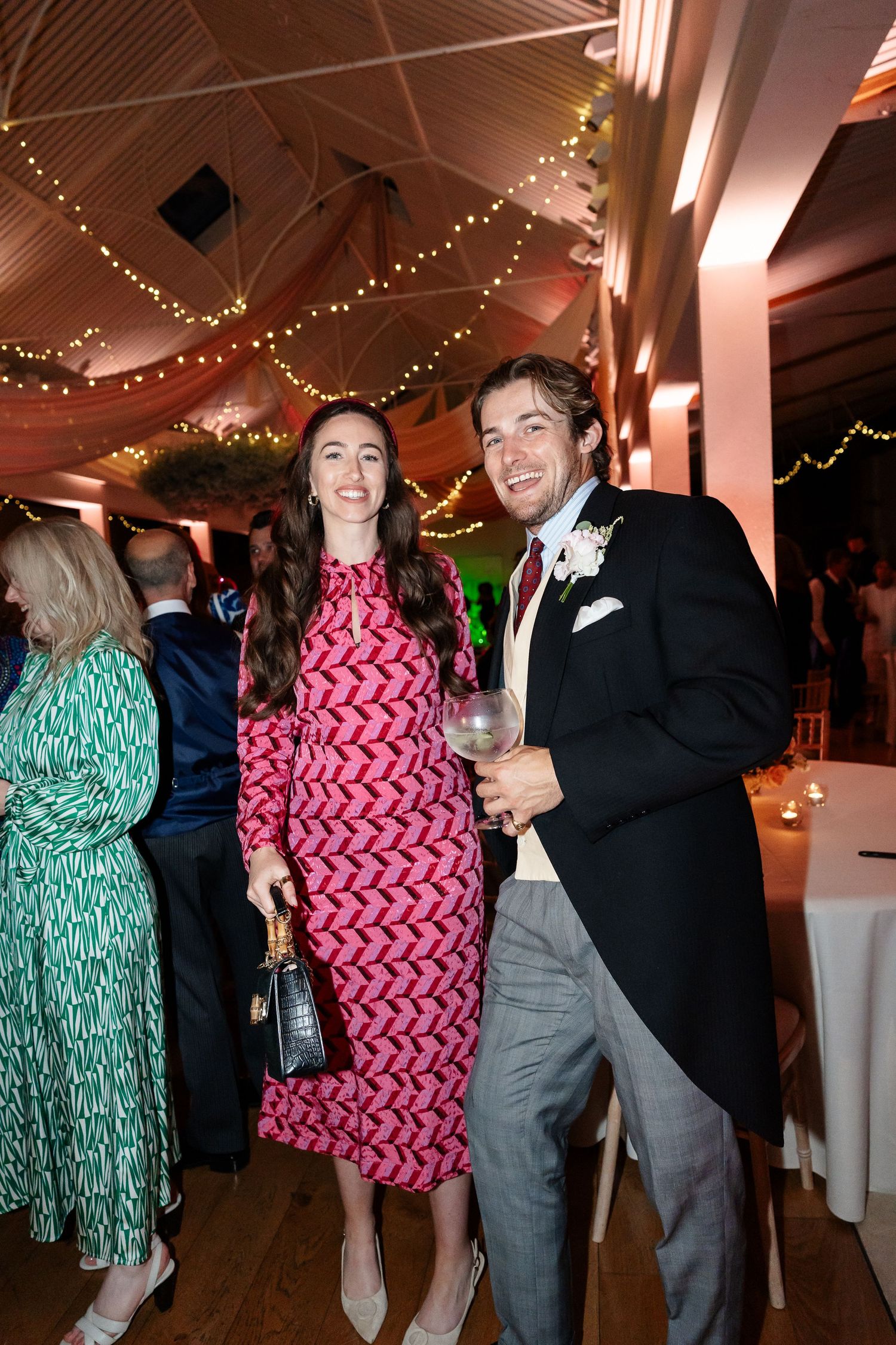 Couple poses smiling at wedding reception under fairy lights, woman in pink patterned dress, man in suit.