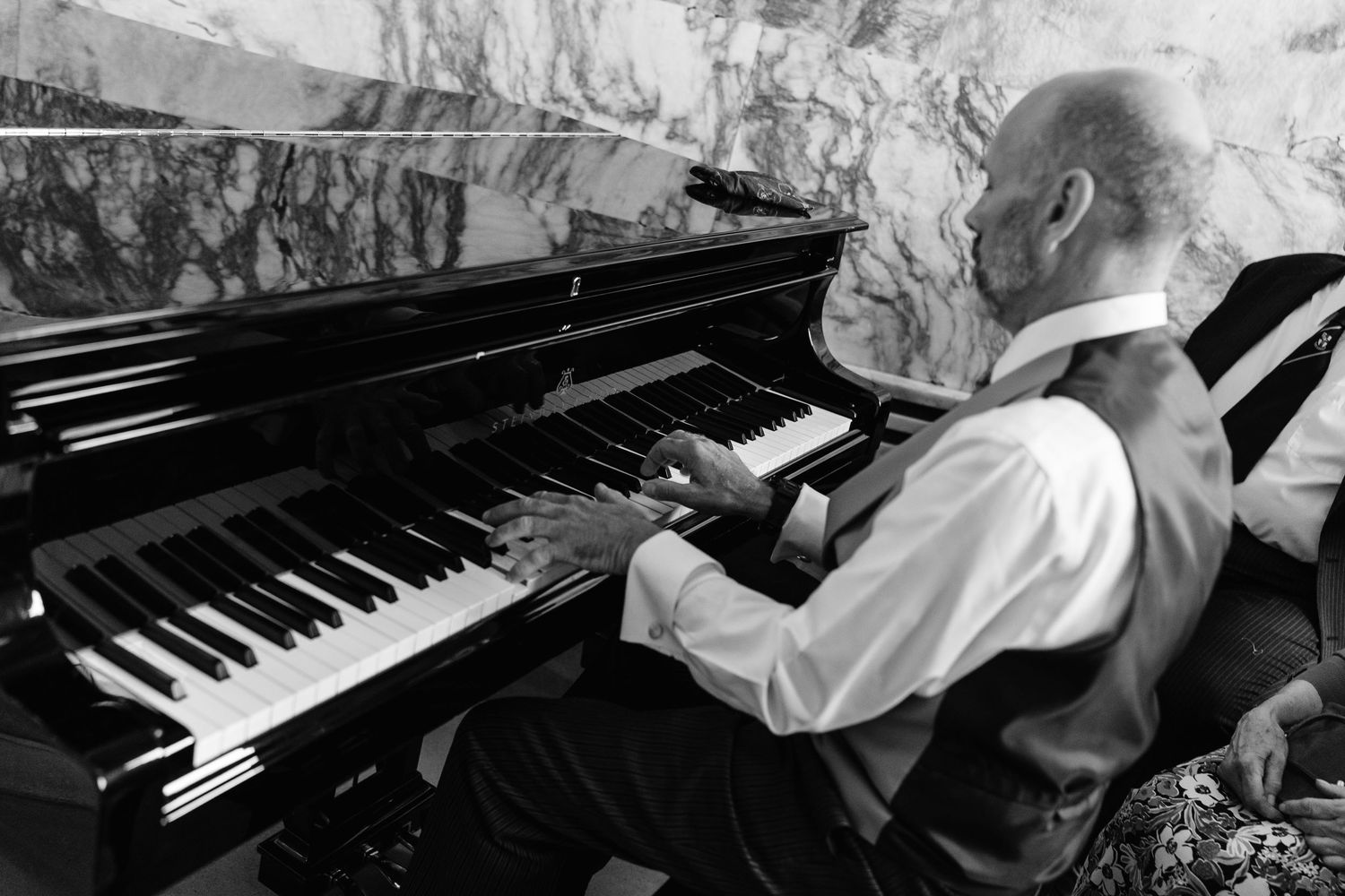Elderly bald man in vest playing grand piano, black and white photography.
