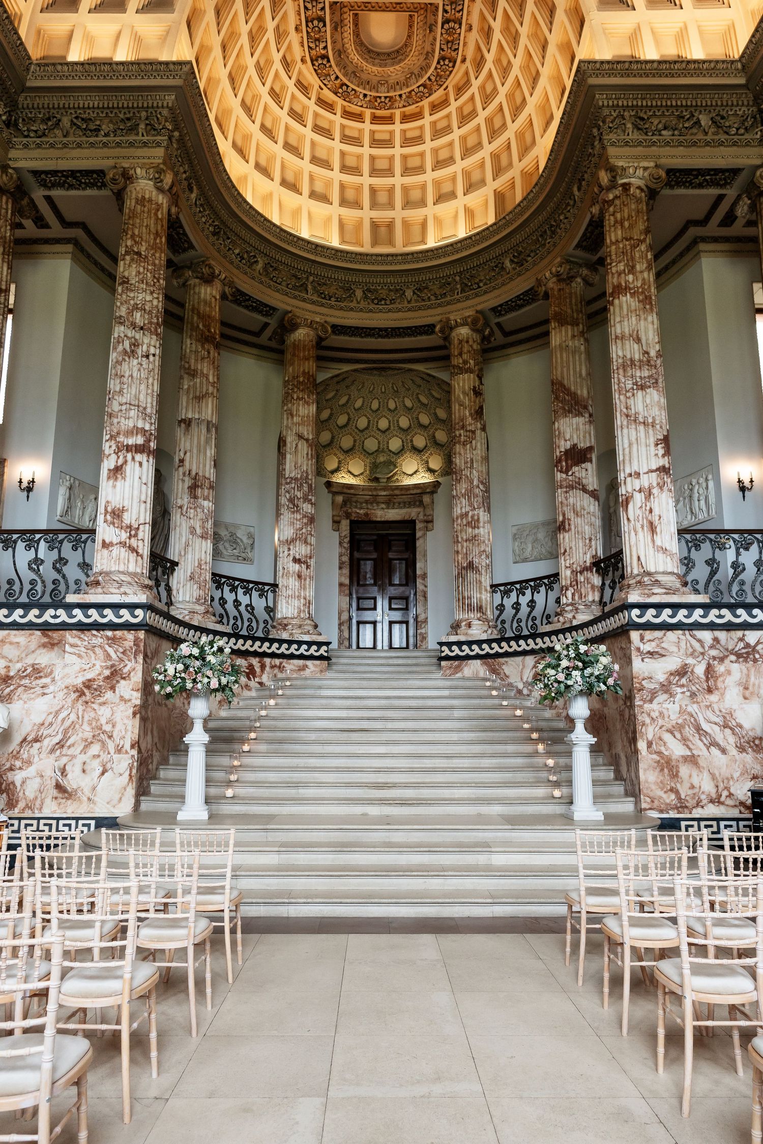 Elegant wedding ceremony setup in a grand neoclassical hall with marble columns, ornate dome ceiling, and floral-decorated staircase.