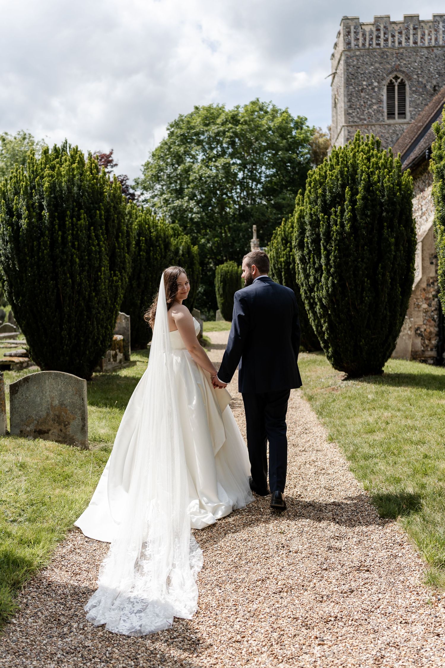 Bride in flowing gown and groom walk hand in hand toward a stone church along a gravel path.
