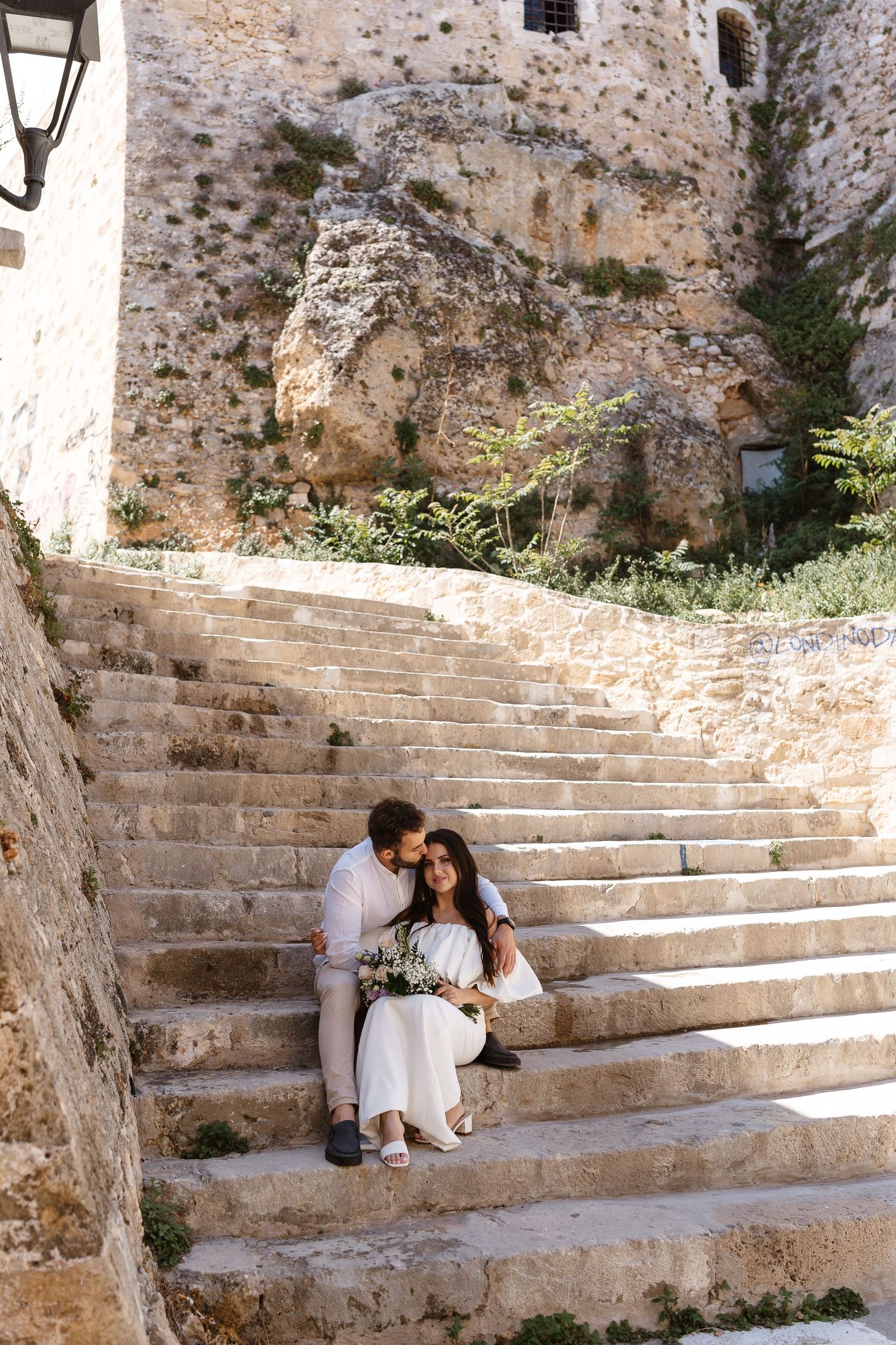 Romantic couple sitting on ancient stone steps embracing near rocky cliff wall with wedding bouquet.