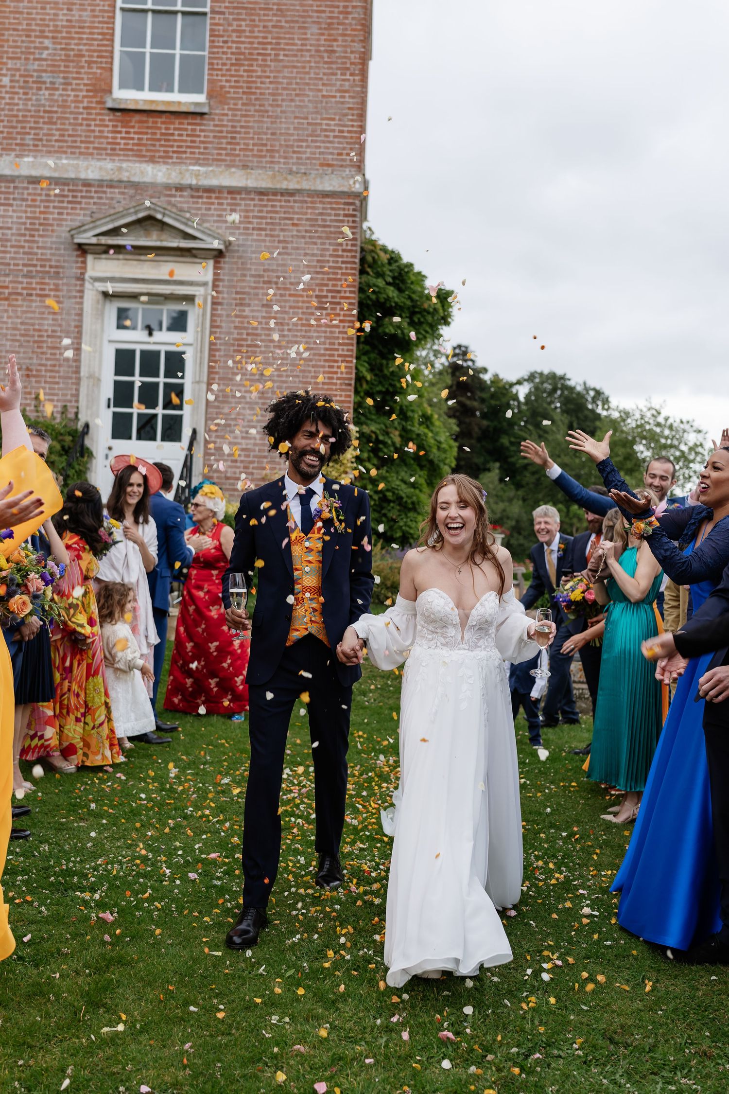 Bride and groom walking through colorful confetti shower thrown by guests outside elegant brick manor house.