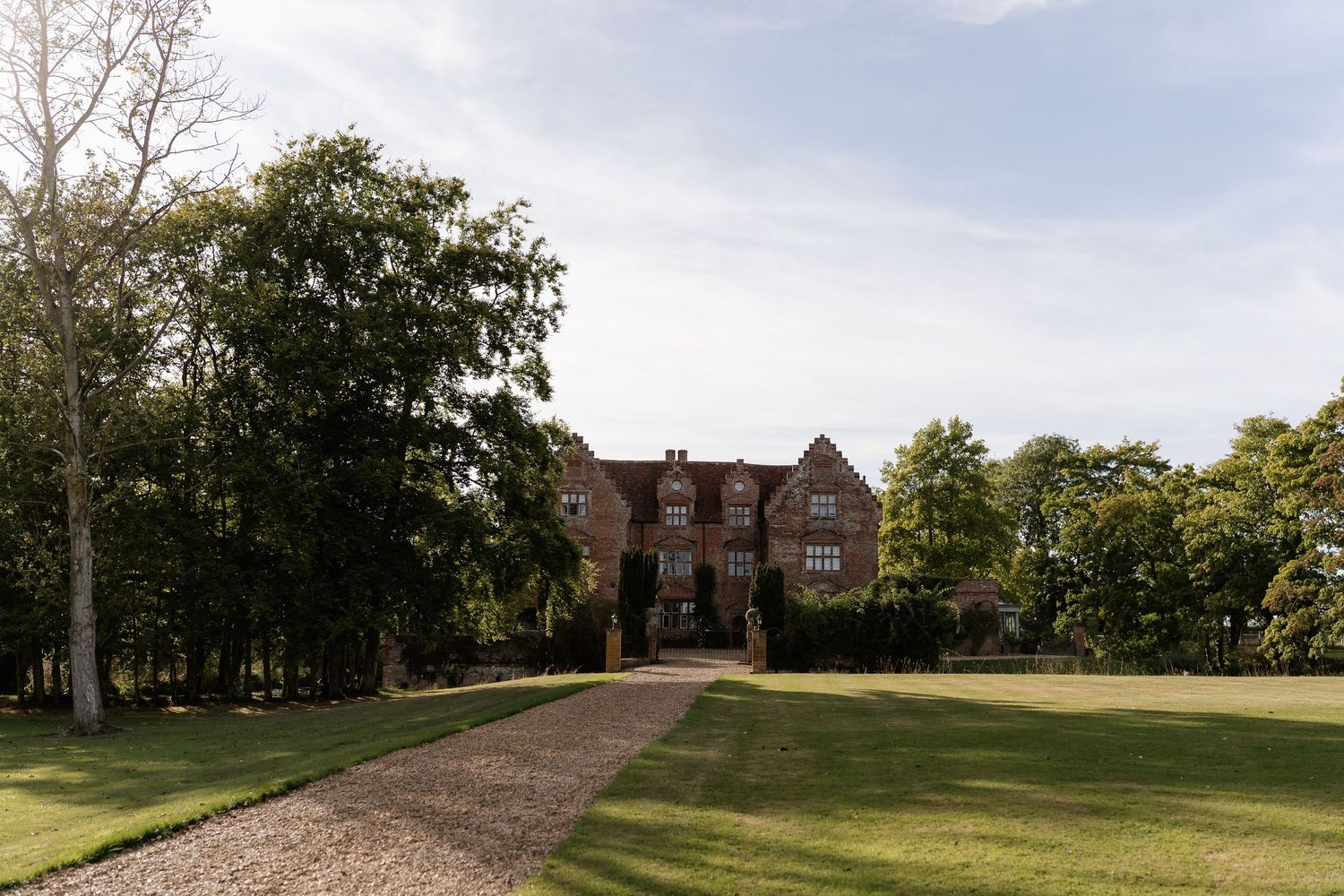 Grand red brick Tudor manor house surrounded by manicured lawns and tall trees on a clear day.