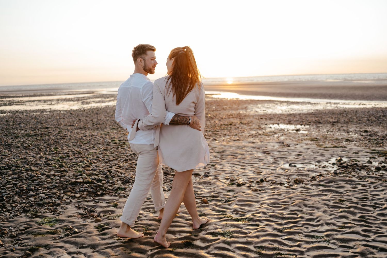 Couple walking barefoot on a sandy beach at sunset, dressed in coordinating white and beige outfits.