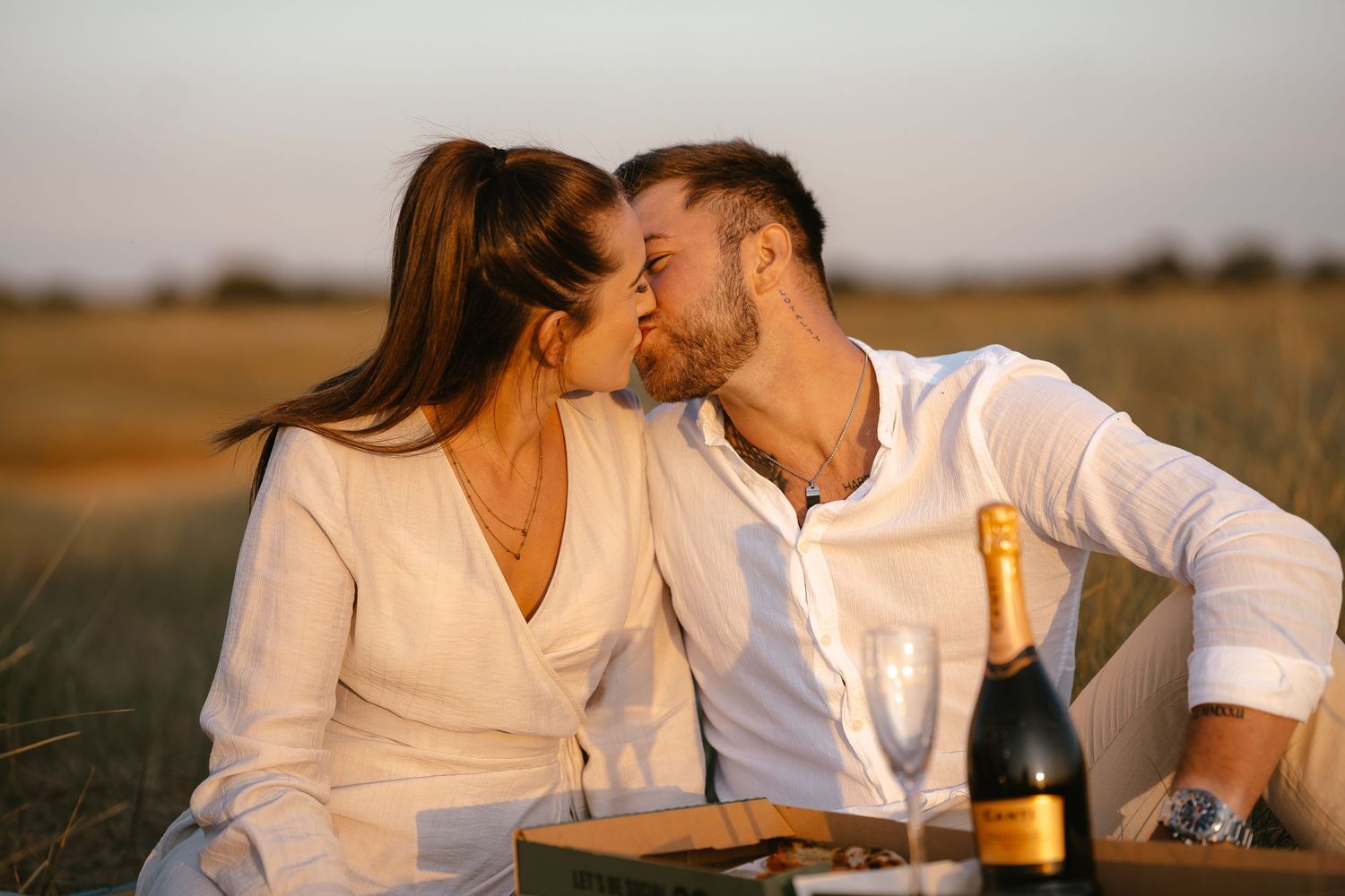 Romantic couple sharing a kiss during a sunset picnic in a field with champagne and glasses nearby.