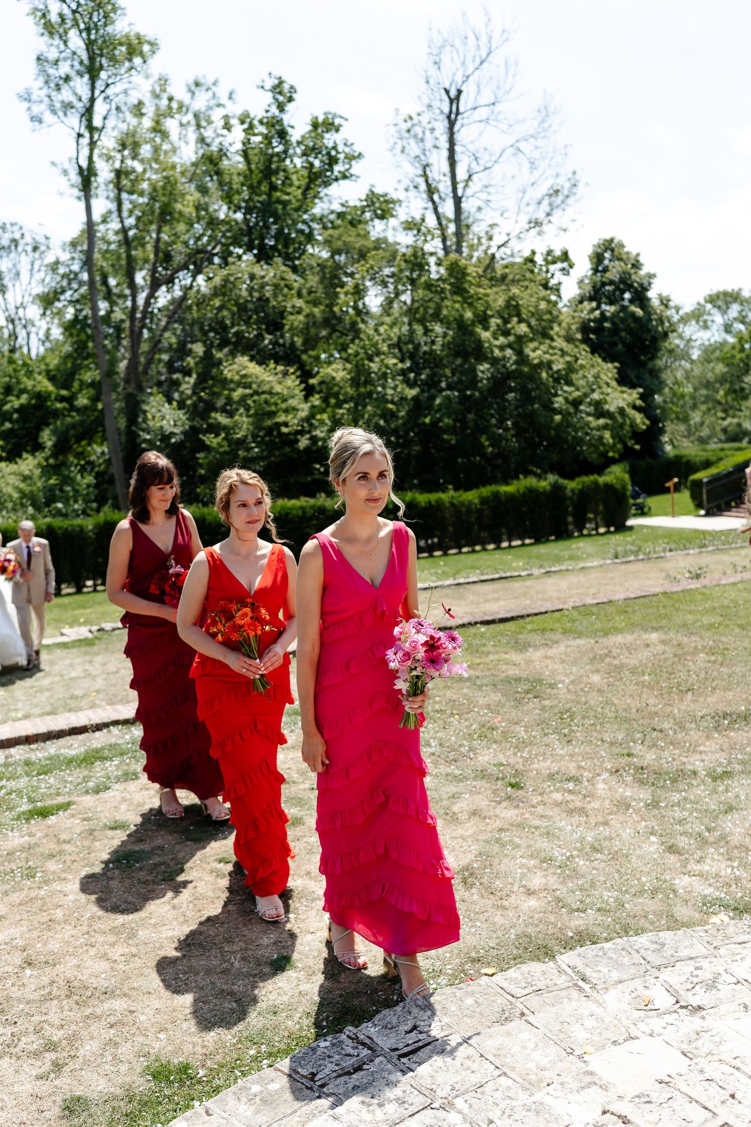 Three bridesmaids in red and pink ruffled dresses walking down outdoor aisle carrying floral bouquets.