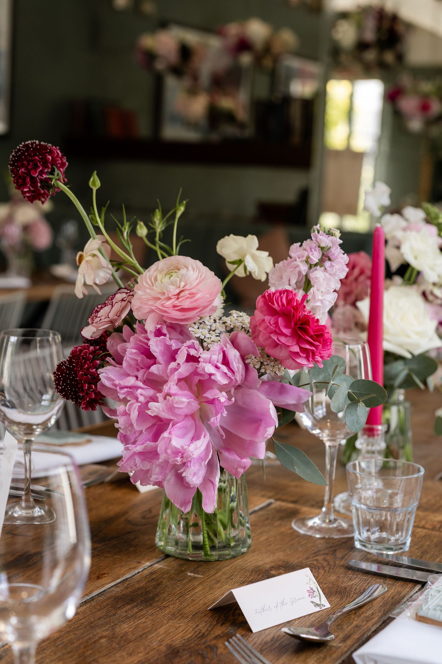 Pink peony and ranunculus wedding centerpiece in glass vase on rustic wooden reception table with crystal glassware.