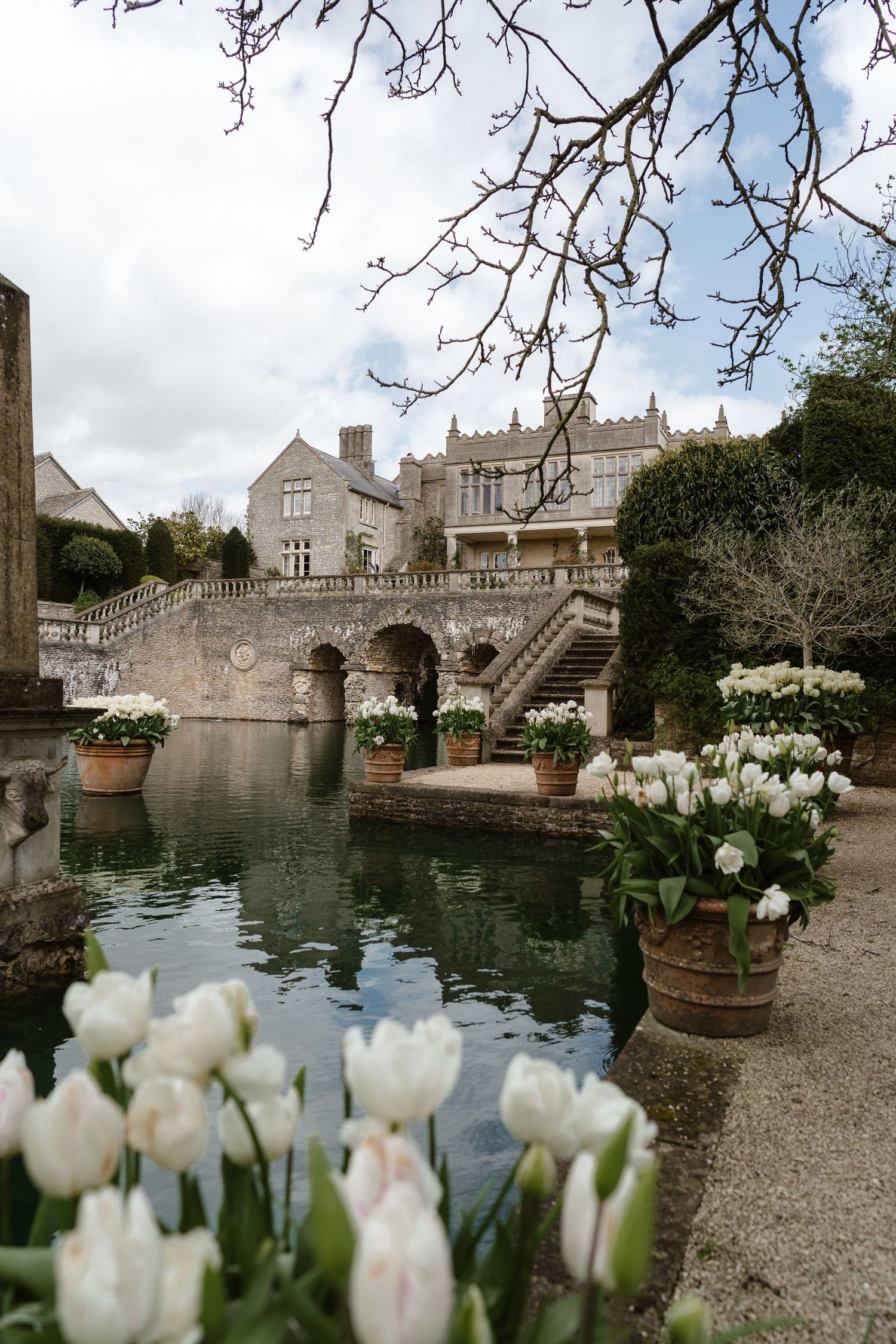Historic stone manor with reflecting pool and white tulips in foreground on cloudy day.