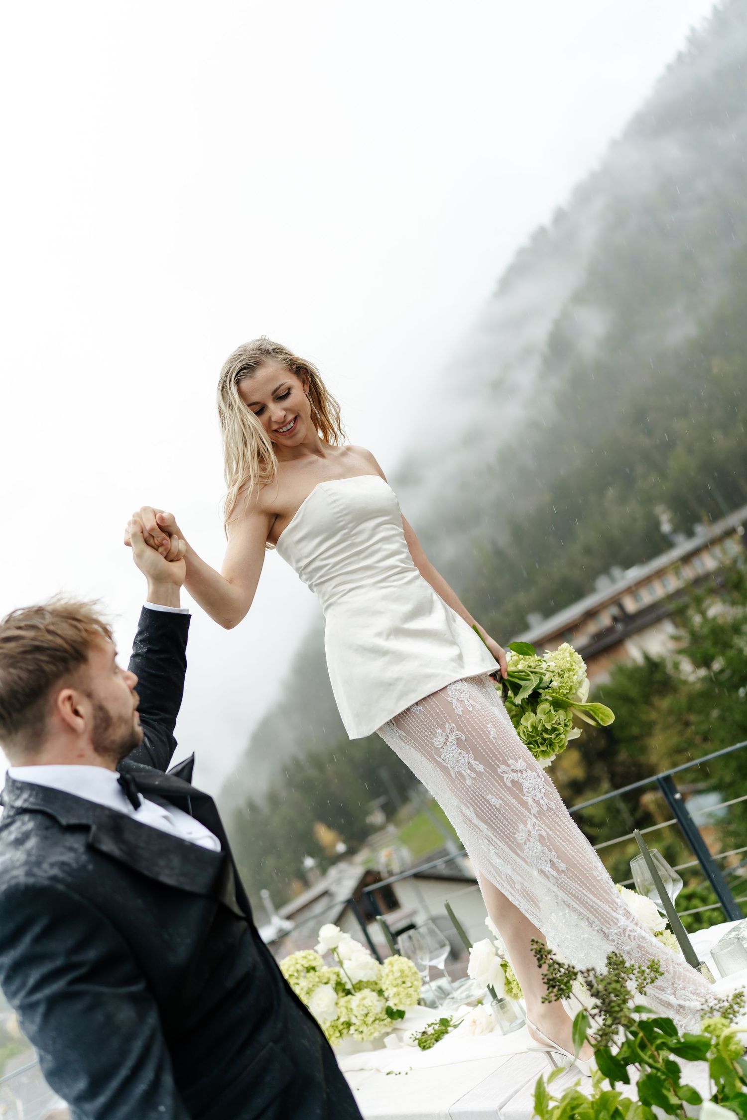 Bride in beautiful white dress dancing on table with groom on terrace in the Dolomites at their romantic and exciting mountain elopement
