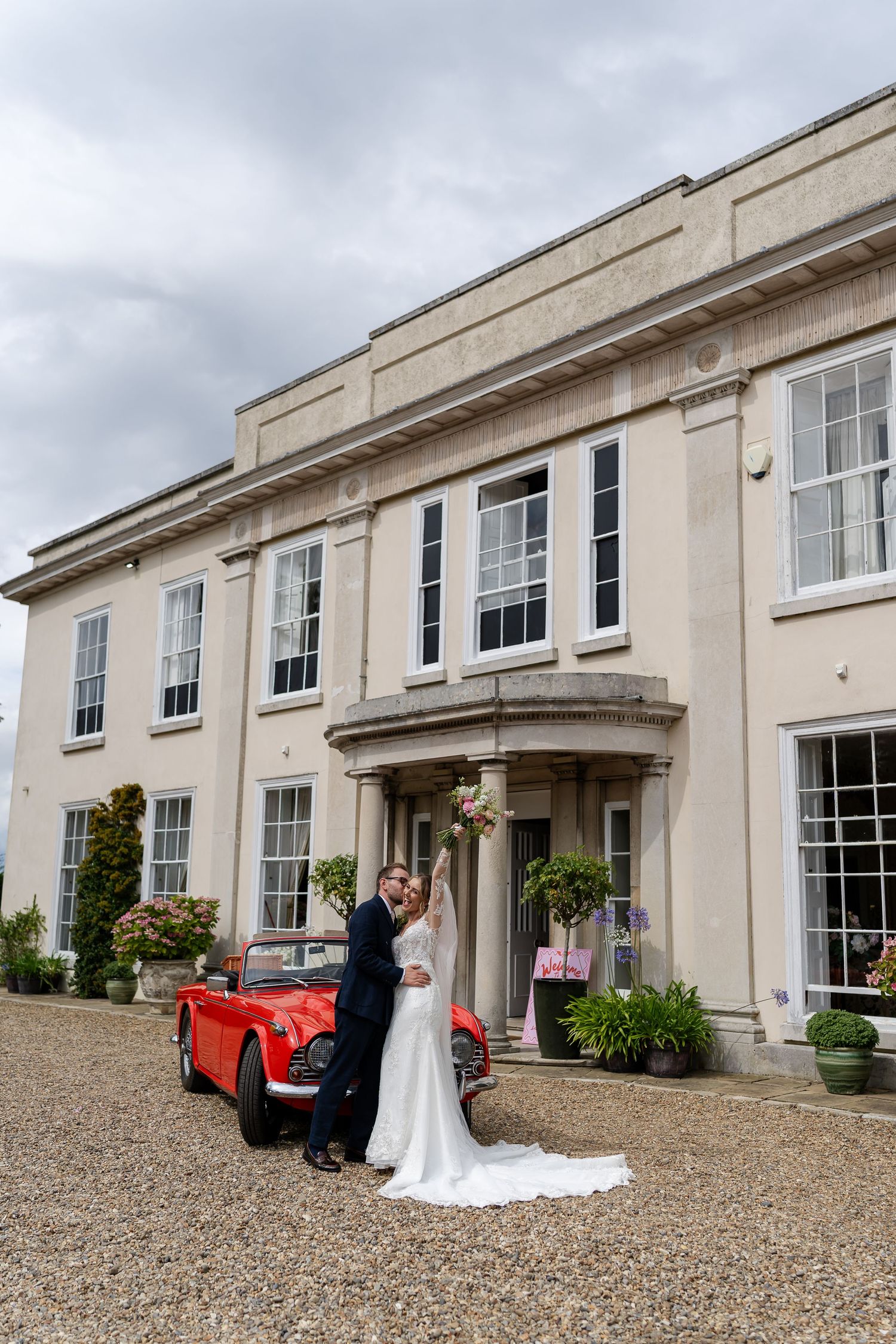Bride and groom embrace beside a classic red convertible in front of a grand Georgian manor house.