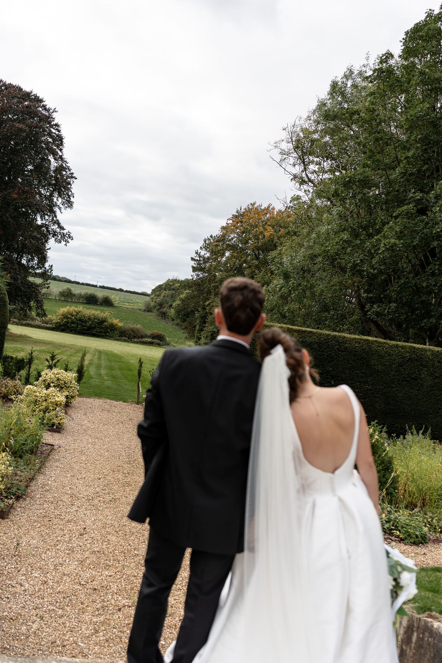 Bride and groom walk hand-in-hand along a garden path surrounded by lush greenery on their wedding day.