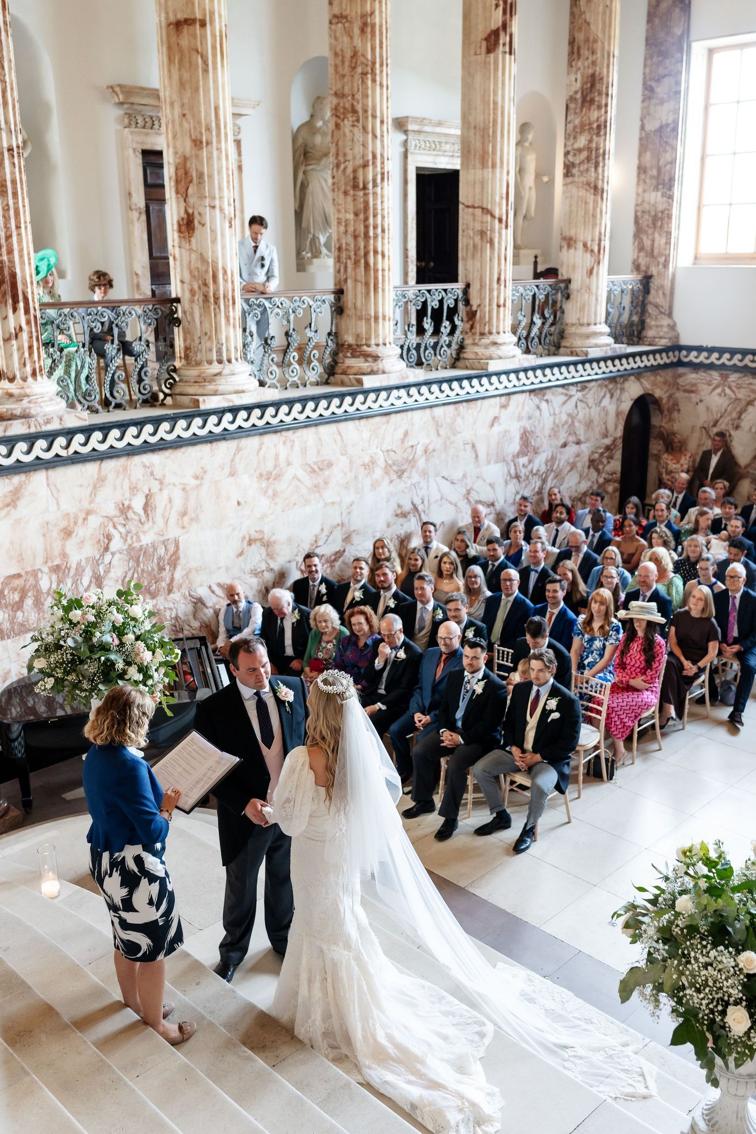 Aerial view of elegant wedding ceremony in ornate marble hall with tall columns and seated guests.