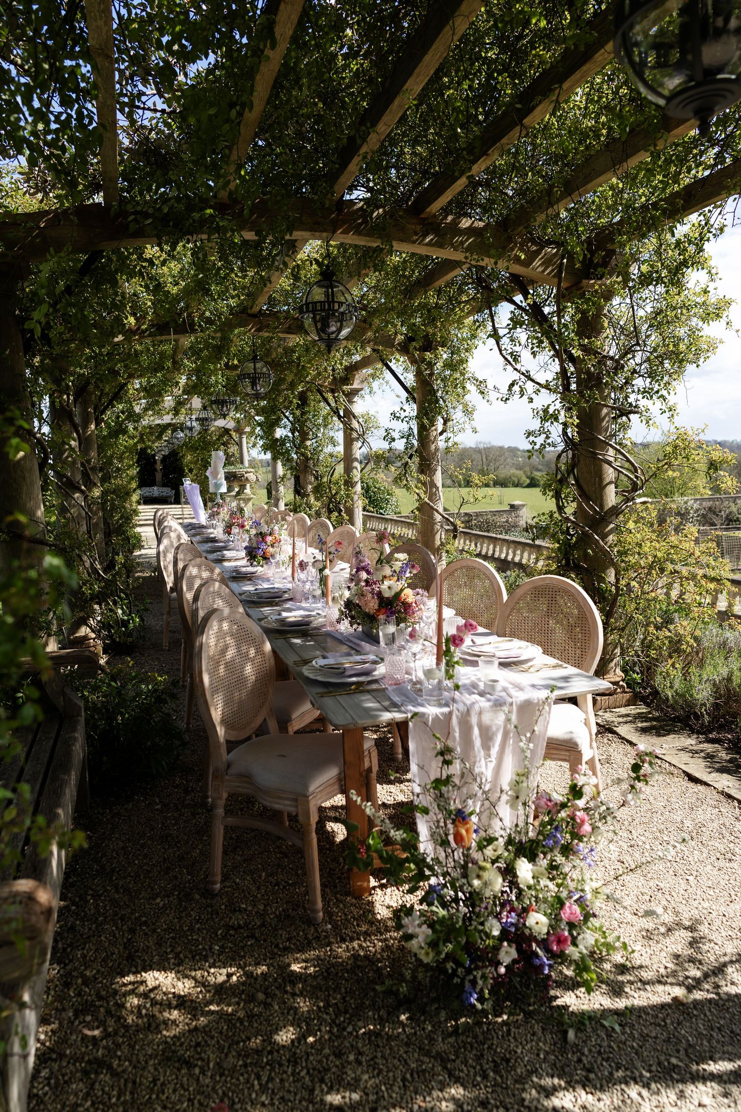 An elegant outdoor wedding reception table set under a green pergola with colorful floral arrangements.