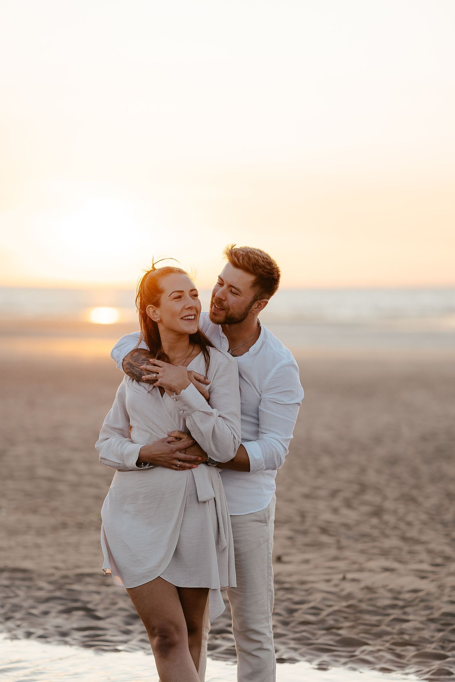 A man tenderly embracing a woman from behind on a beach at sunset, both wearing white outfits.
