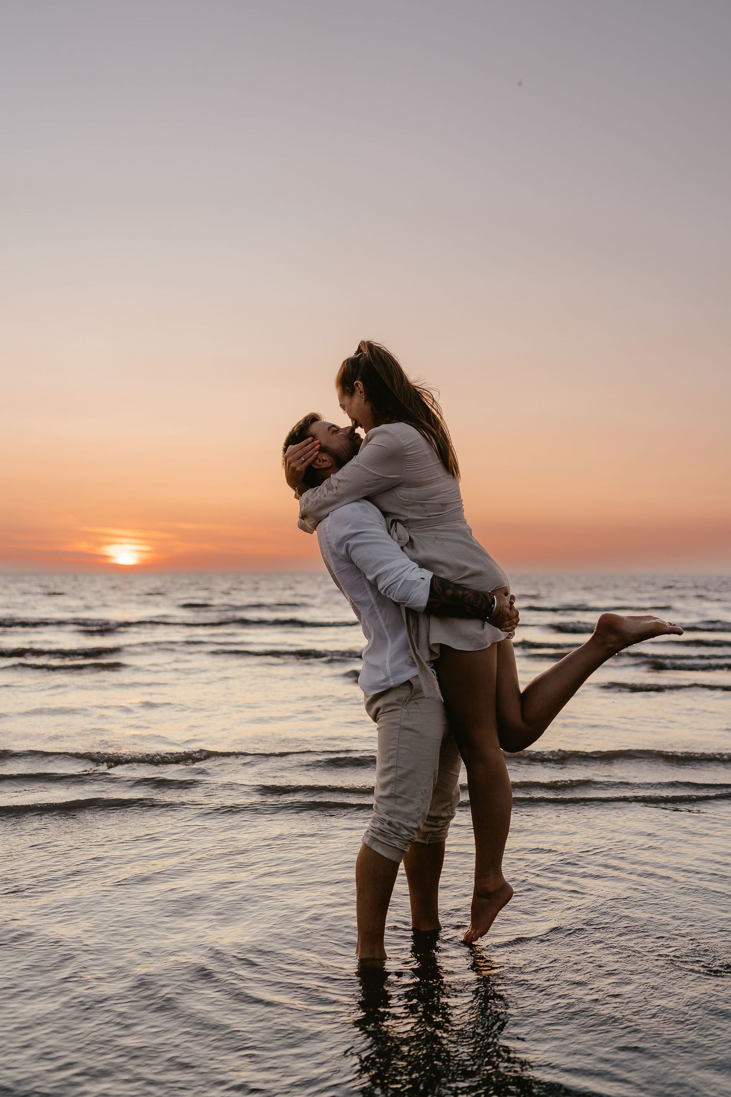 Man lifting woman in romantic embrace while standing in ocean at sunset.