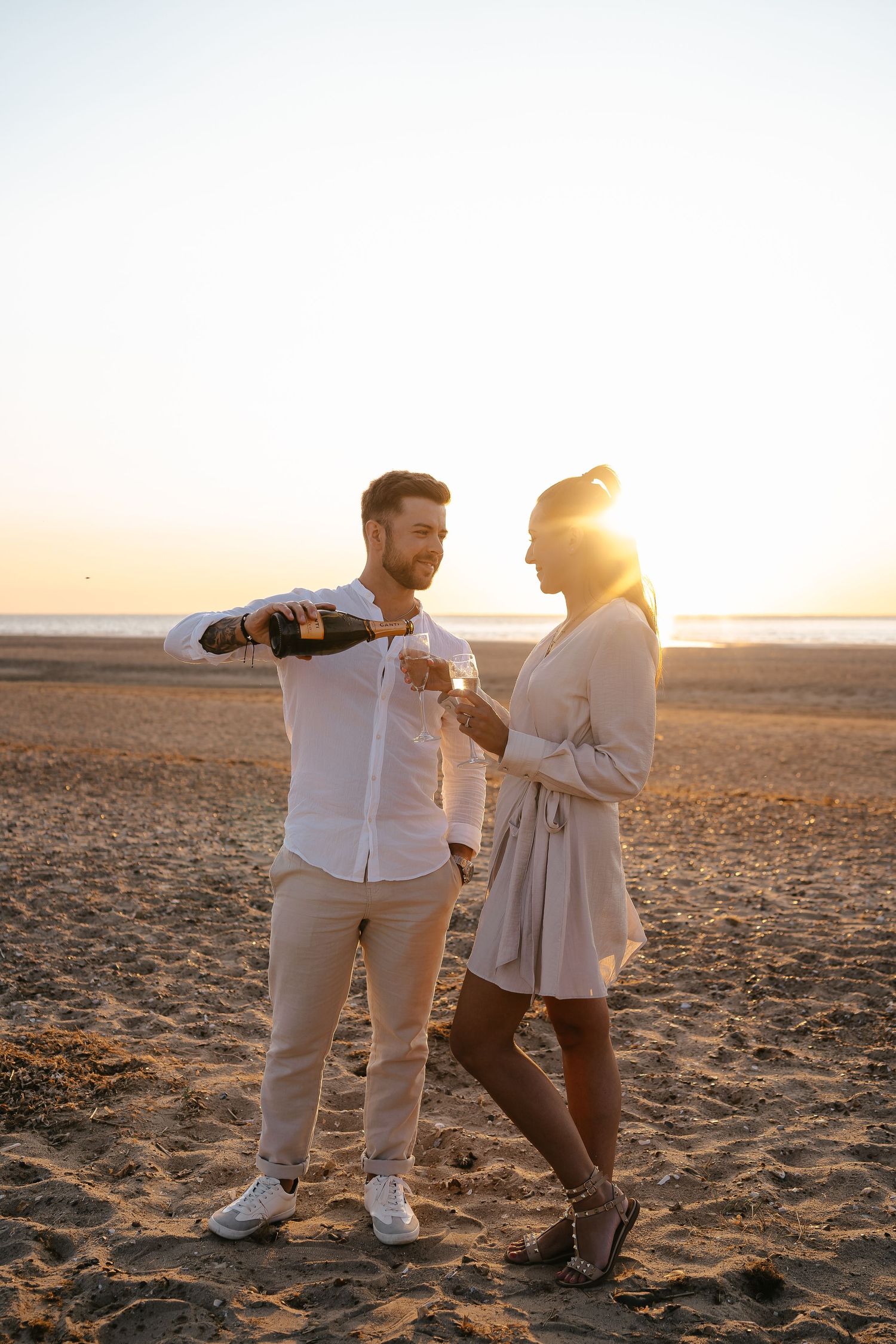 Couple sharing champagne on a sandy beach at golden sunset, man pouring wine into woman's glass.