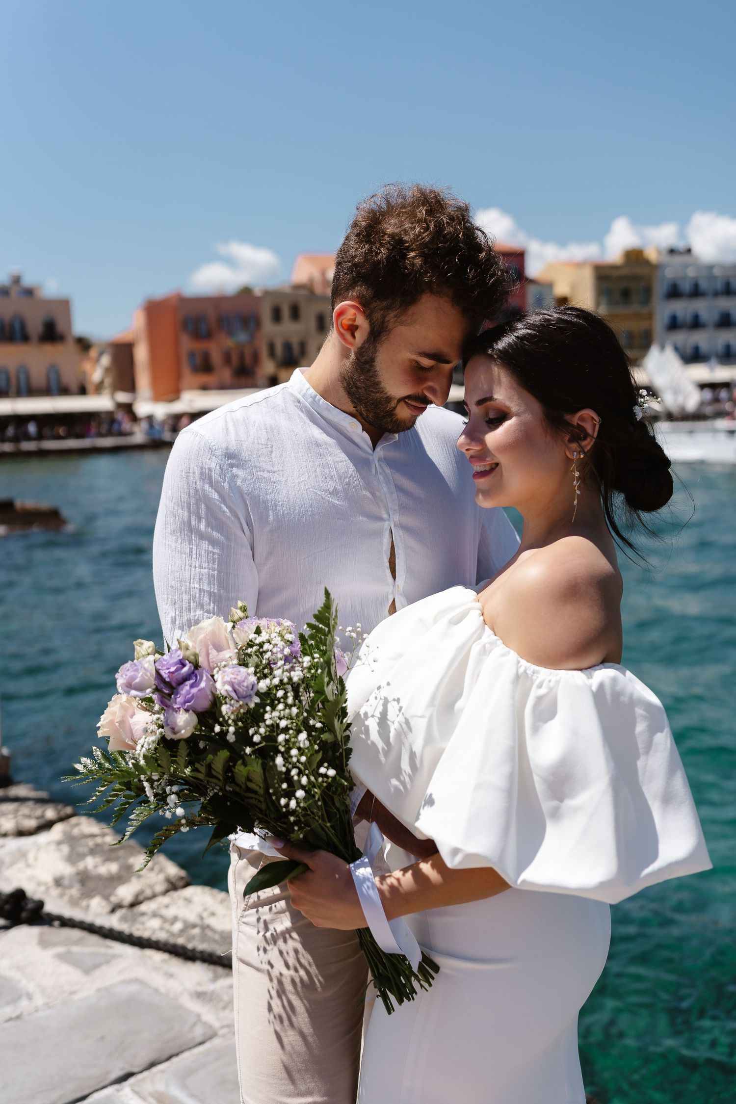 Groom tenderly embraces bride holding a purple and white bouquet on a sun-drenched Greek waterfront promenade.