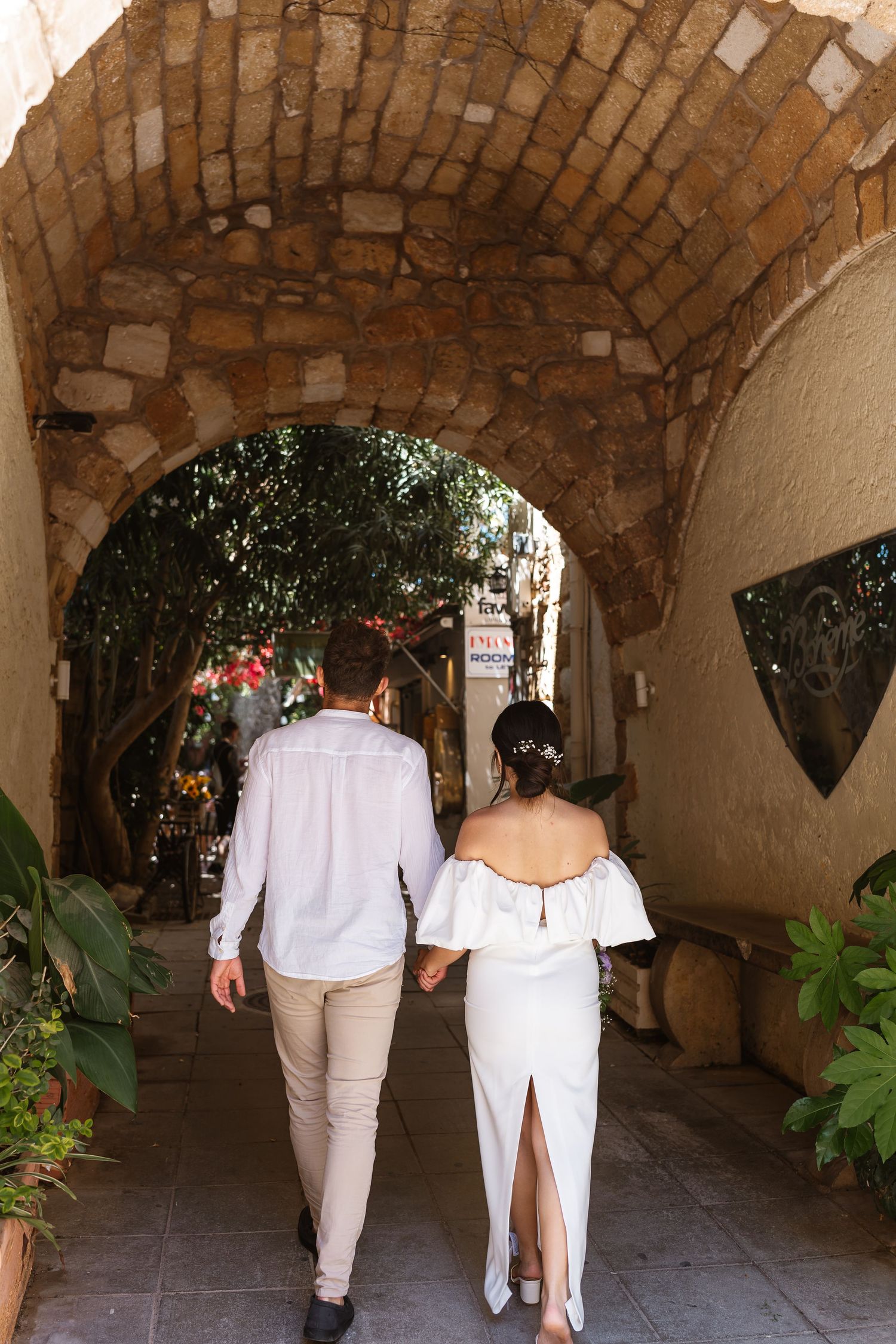 Bride and groom walking hand-in-hand through a rustic stone archway into a lush Mediterranean alleyway.