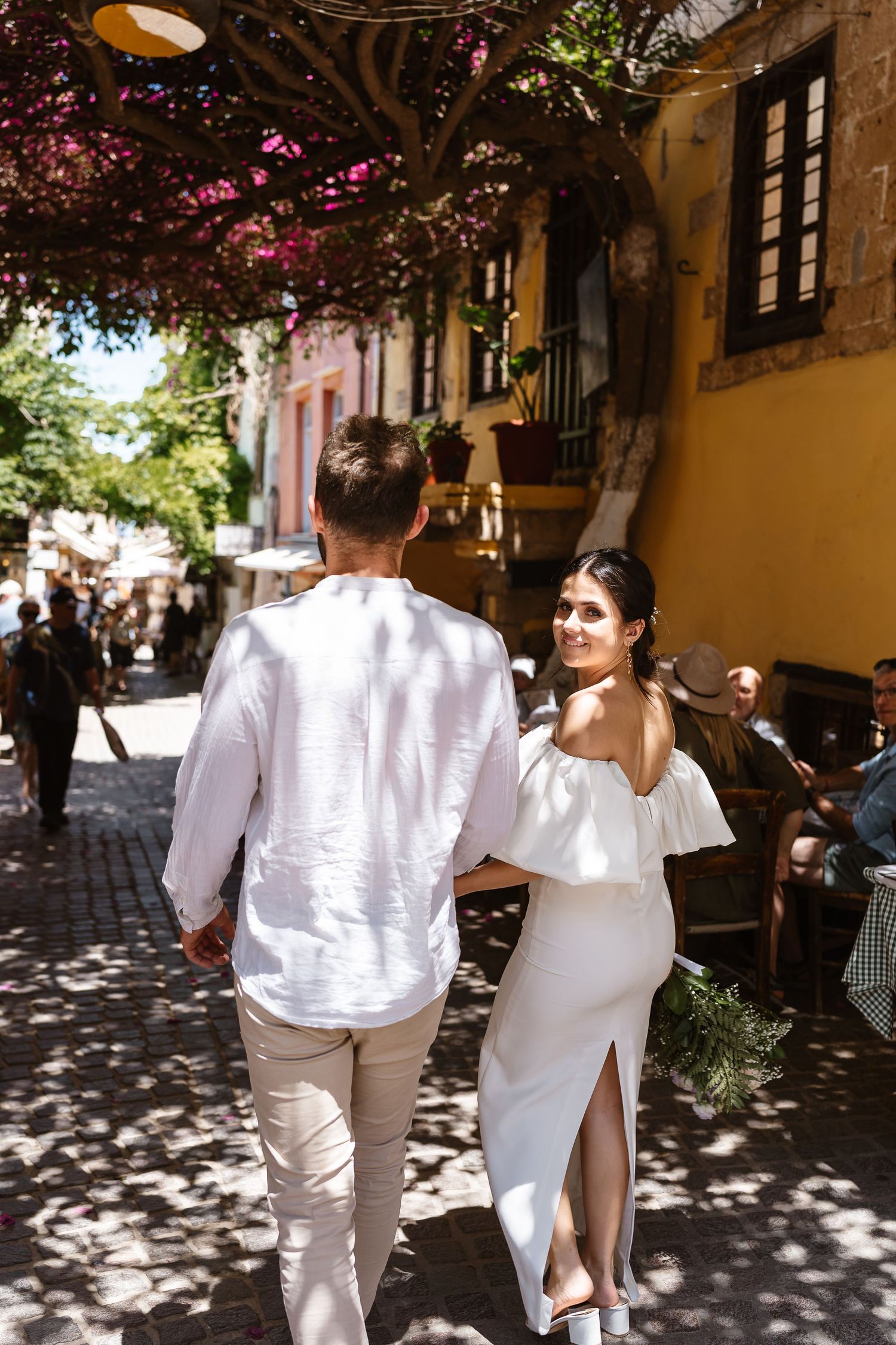 Bride in white off-shoulder dress holds groom's hand walking through a charming Mediterranean cobblestone street.