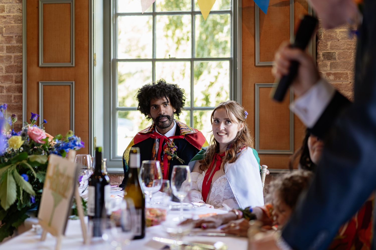 Groom in decorative jacket and bride with red ribbon sit together at wedding reception table listening to speeches.