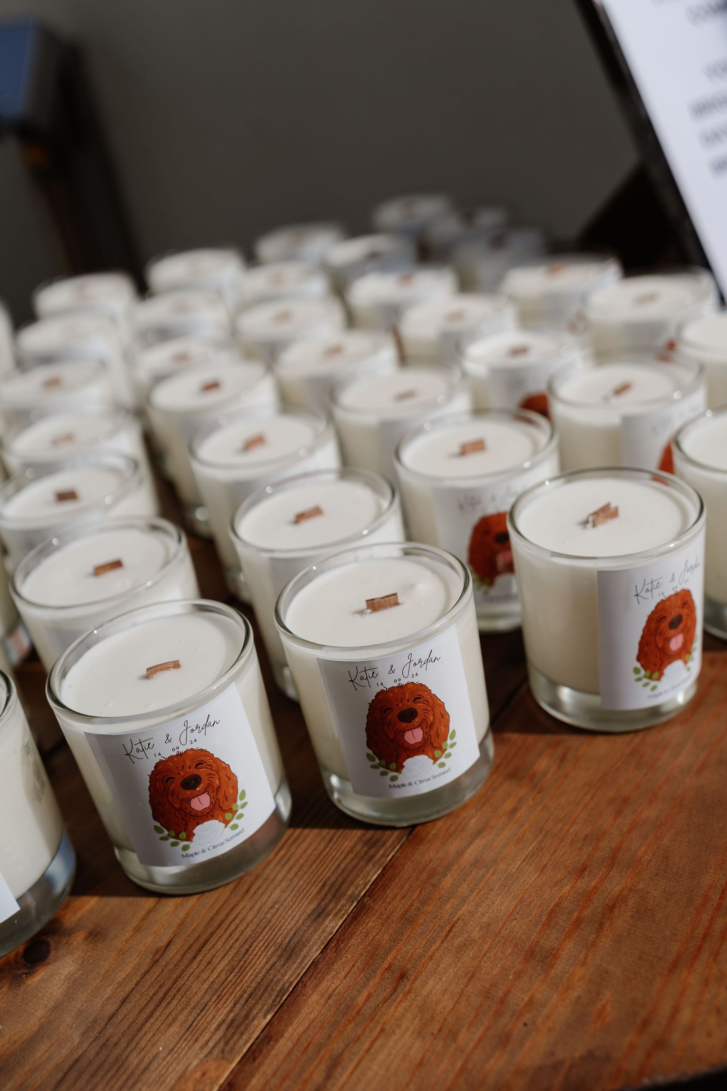 Rows of white candle wedding favors with illustrated dog labels displayed on a wooden table at a reception.