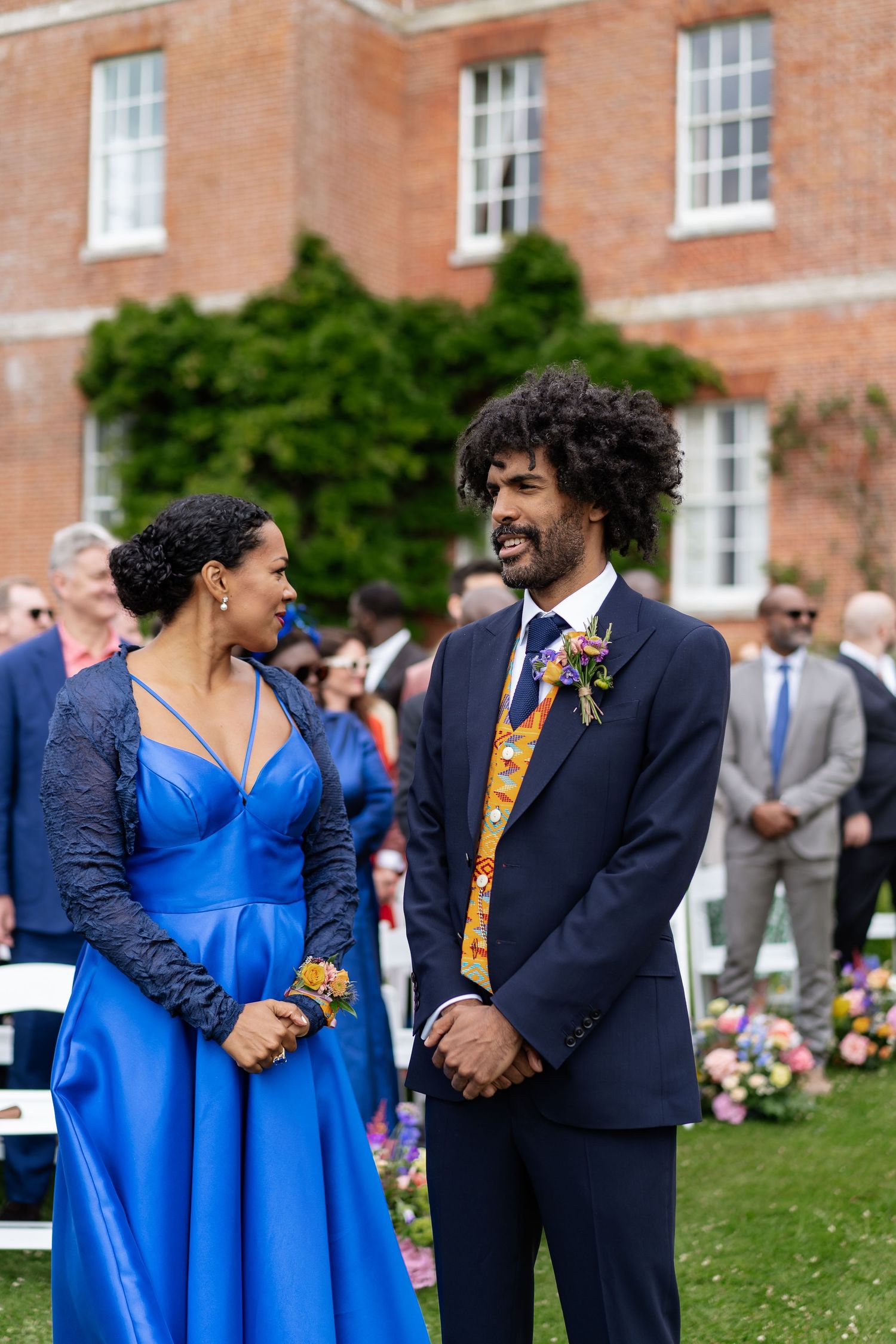 Groom in navy suit with floral waistcoat smiling at woman in blue gown at outdoor wedding ceremony.