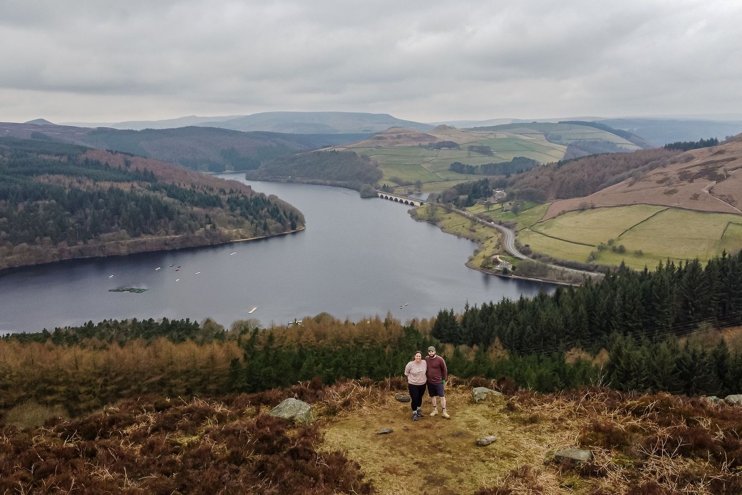 Two hikers overlooking a scenic reservoir surrounded by forested hills and rolling valleys.