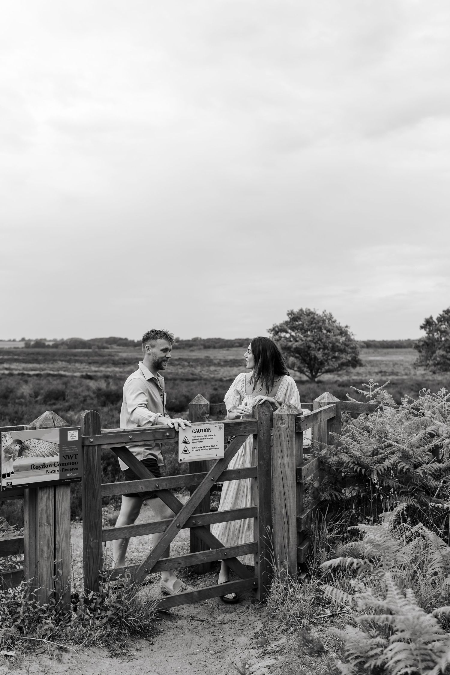 Couple leans on a rustic wooden gate surrounded by ferns in a black and white countryside engagement photo.
