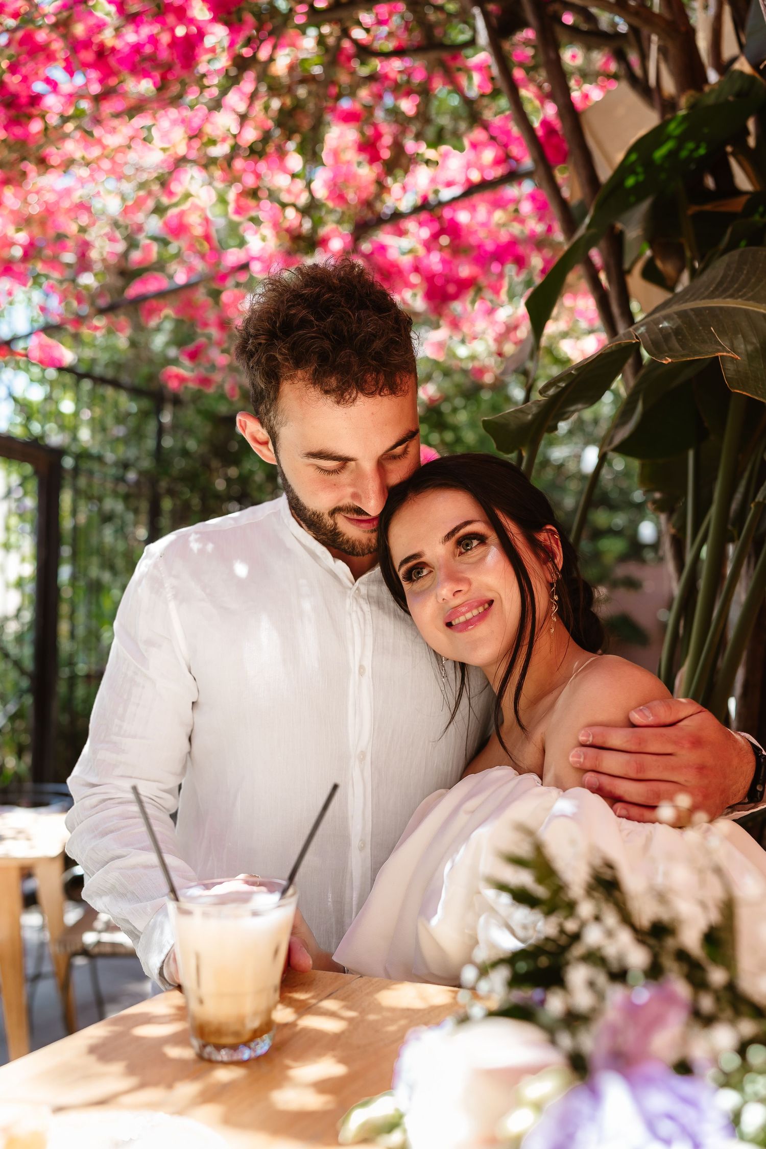 Romantic couple embracing at a cafe table under vibrant pink bougainvillea flowers with iced coffee drinks.