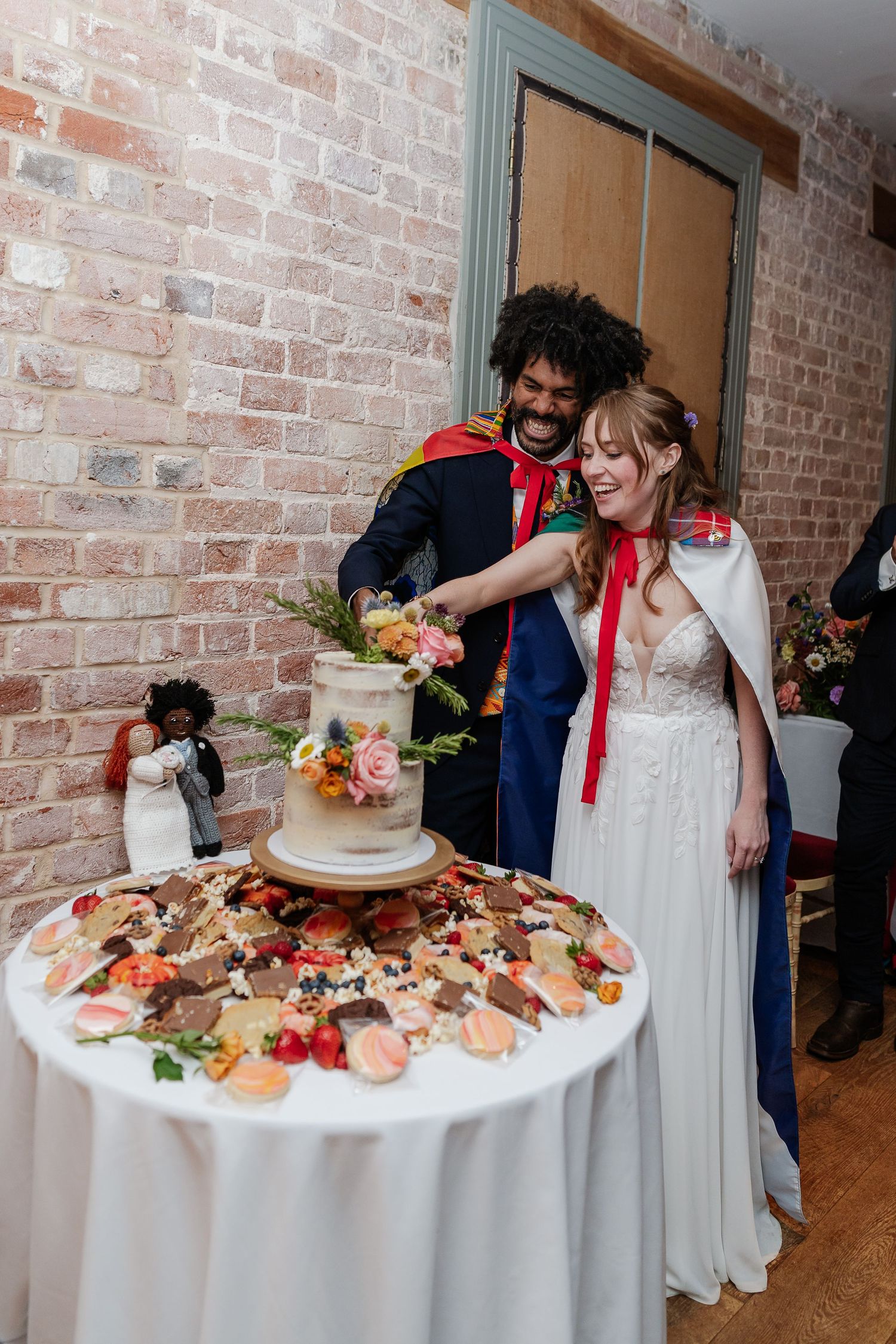 Newlyweds cut wedding cake decorated with flowers at rustic brick wall venue.